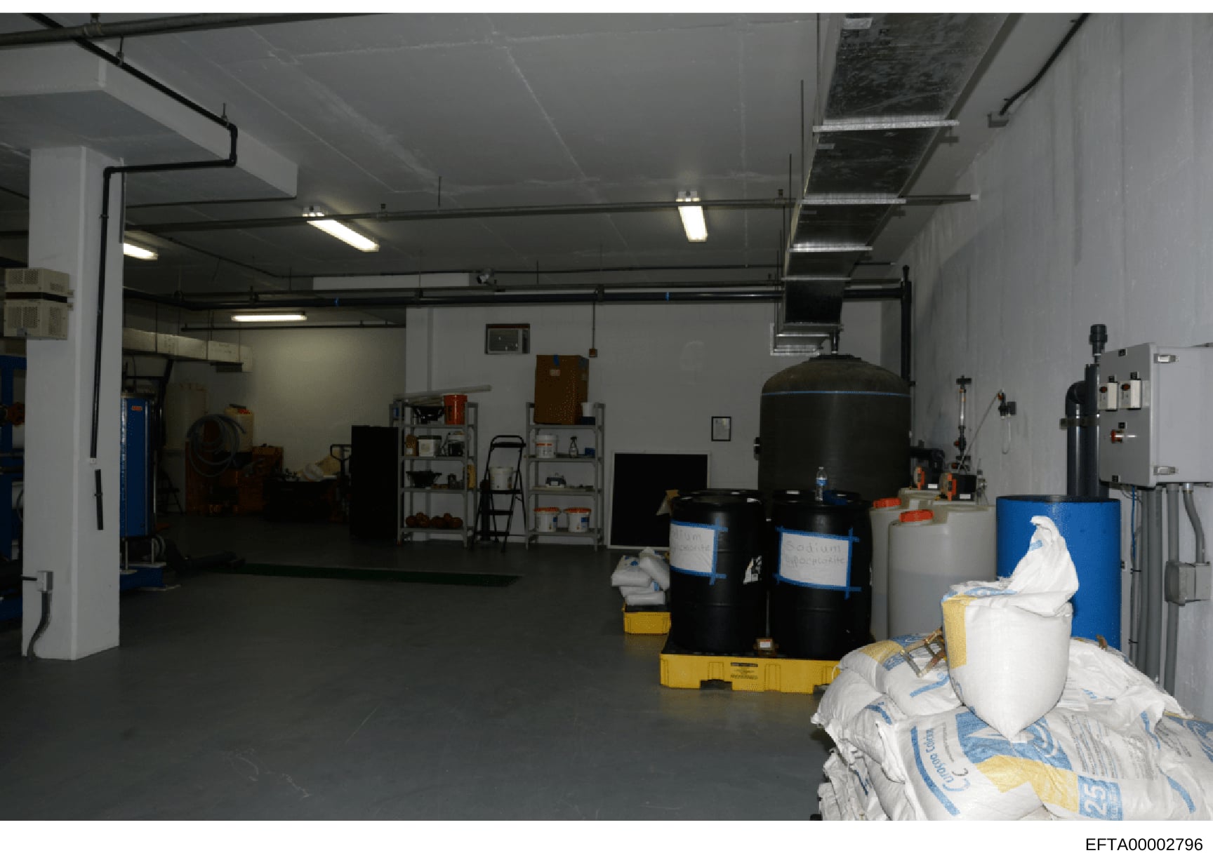 This is a photograph of a large, warehouse-like industrial space with chemical storage and laboratory equipment. The room contains multiple large cylindrical tanks, chemical drums (black and white), storage shelves, and various industrial equipment. 