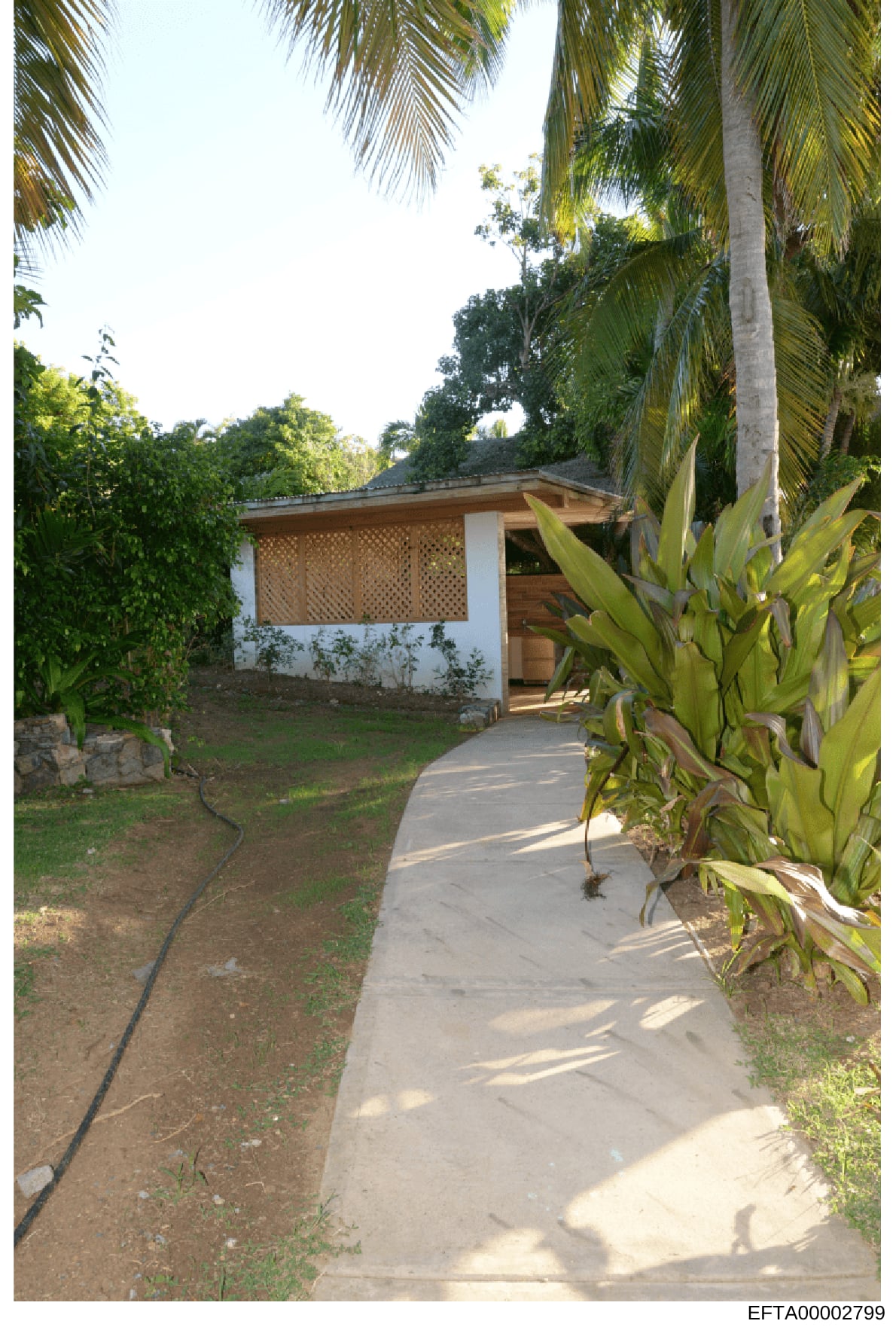 This photograph shows a small white building with latticed wooden panels and a covered entrance, situated within a tropical garden environment. The structure appears to be a guest house, storage facility, or ancillary building on a larger property, s