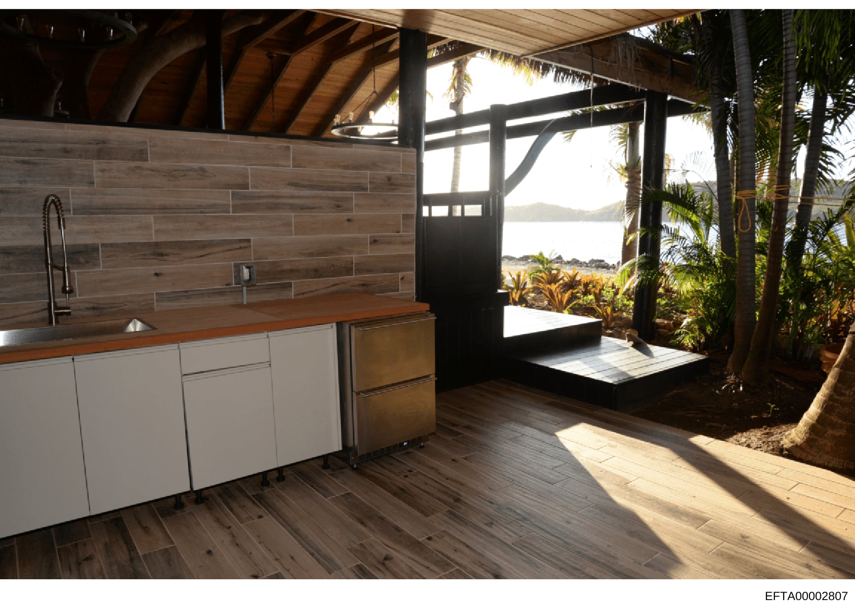 This is a photograph of a modern kitchen and open-air living area in a tropical beachfront property. The image shows a contemporary kitchen with white cabinetry and wood countertops, adjacent to a covered outdoor pavilion with views of water and dist