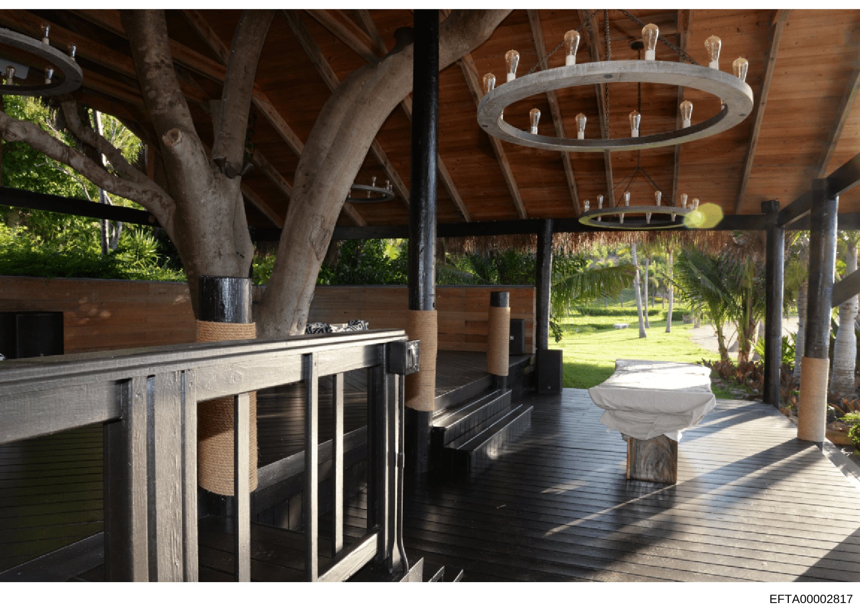 This photograph shows the interior of a covered outdoor pavilion or entertainment structure with exposed wooden beams, a kitchen counter with bar seating on the left, decorative circular light fixtures hanging from the ceiling, and views of landscape