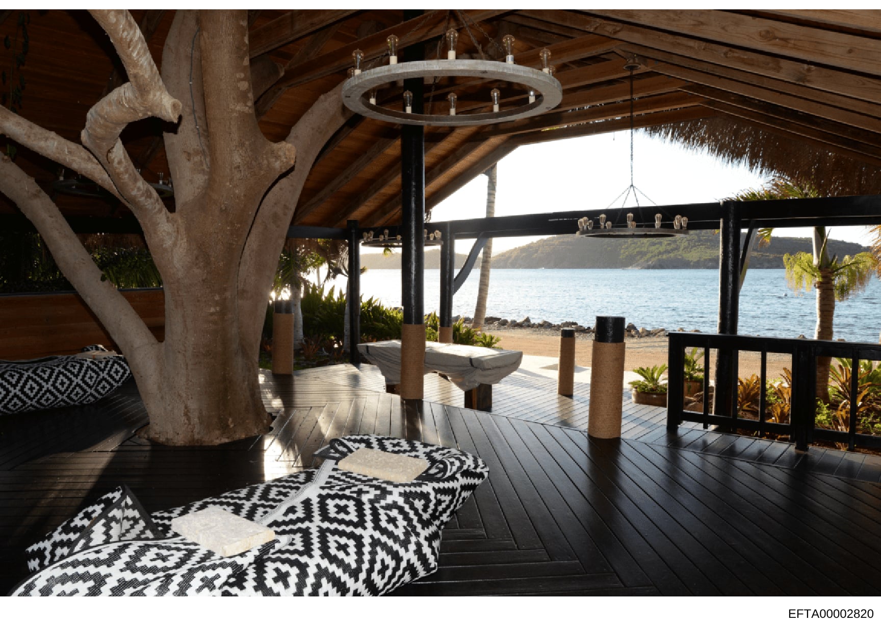This is a photograph of an upscale beachfront property's covered outdoor deck or patio area. The image shows a modern, luxuriously appointed space with high-end finishes including a large circular chandelier, natural wood beam structural elements, de