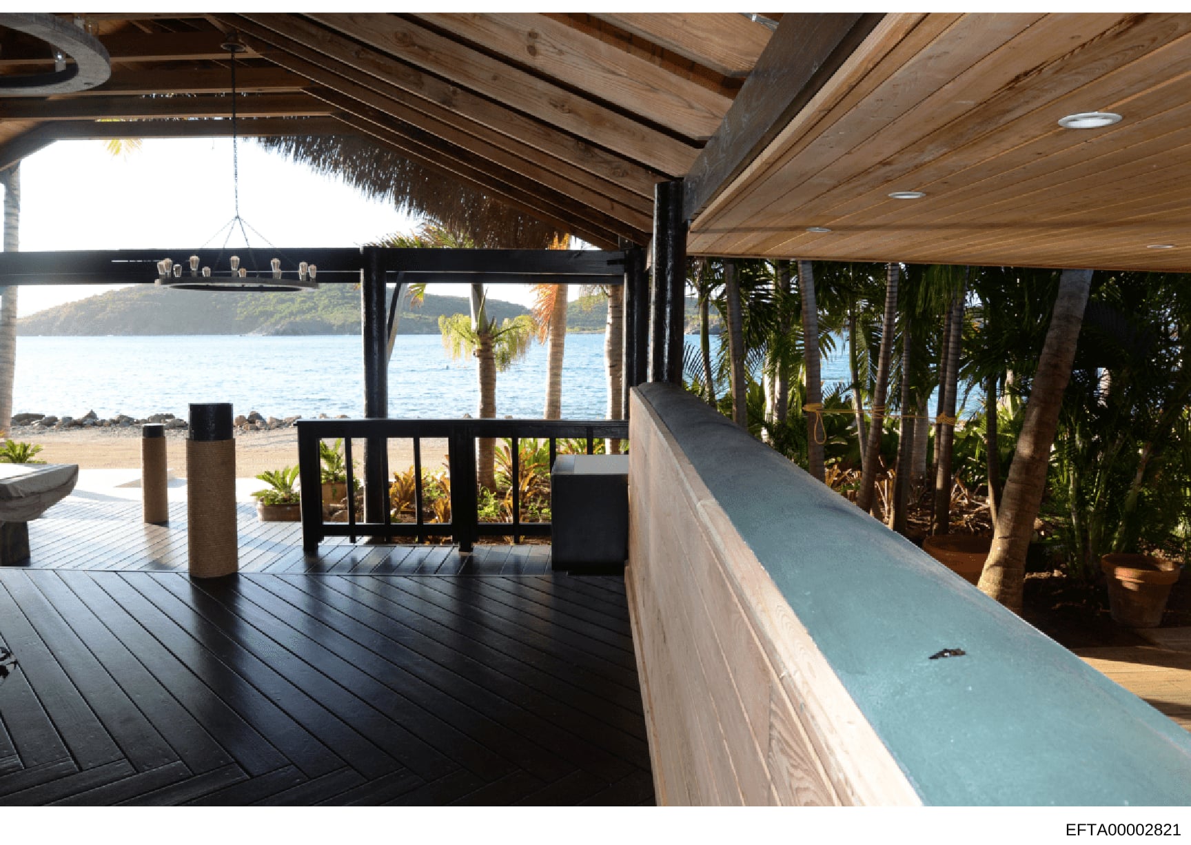 This is a photograph of a luxury beachfront property's interior deck and patio area. The image shows a covered wooden structure with expansive ocean views, tropical landscaping with palm trees, modern architectural design including wooden beam ceilin