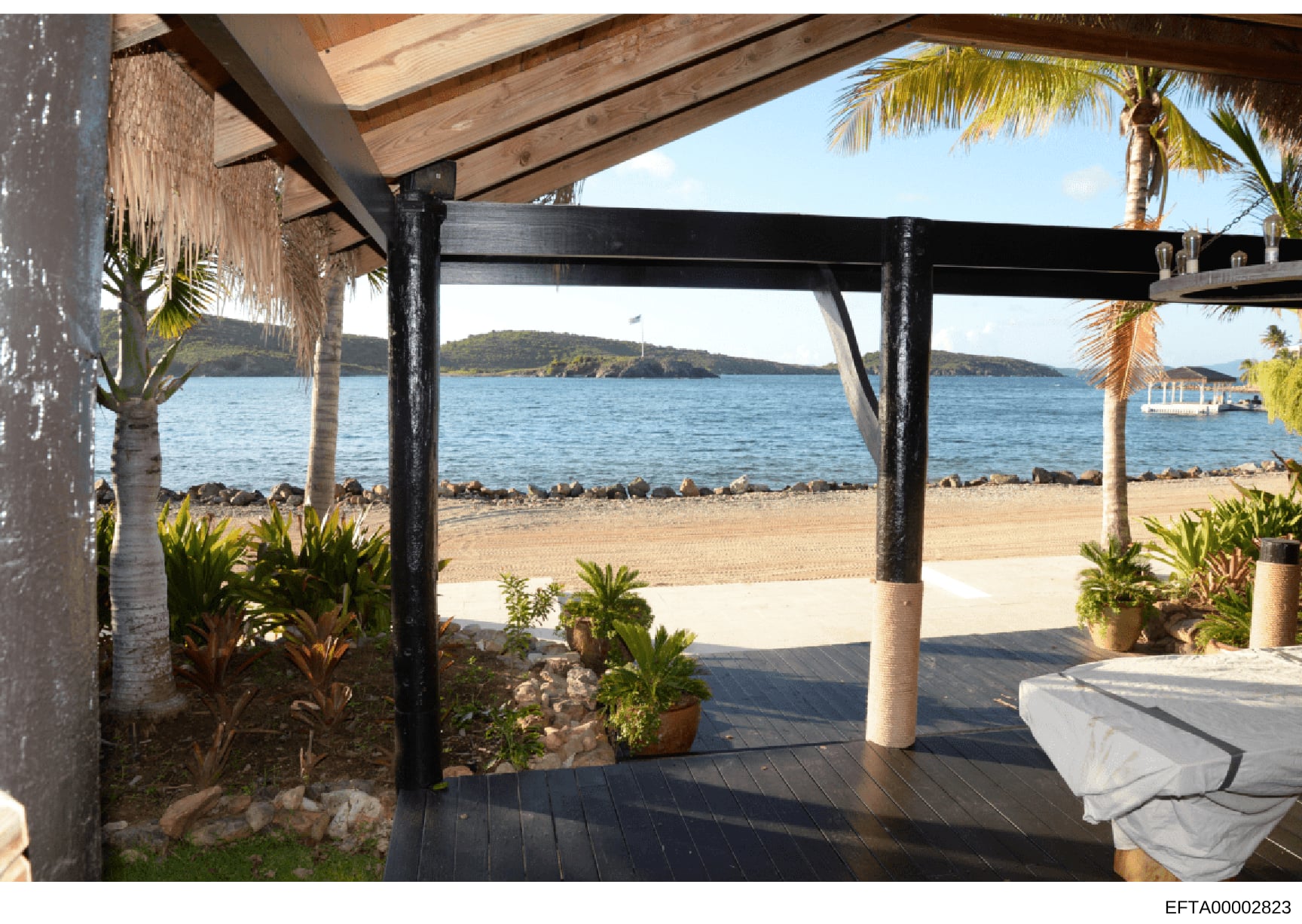 This photograph shows a covered outdoor deck or pavilion structure at a beachfront property, featuring views of a sandy beach, ocean, and distant islands. The structure has wooden beam construction with black metal supports and includes tropical land