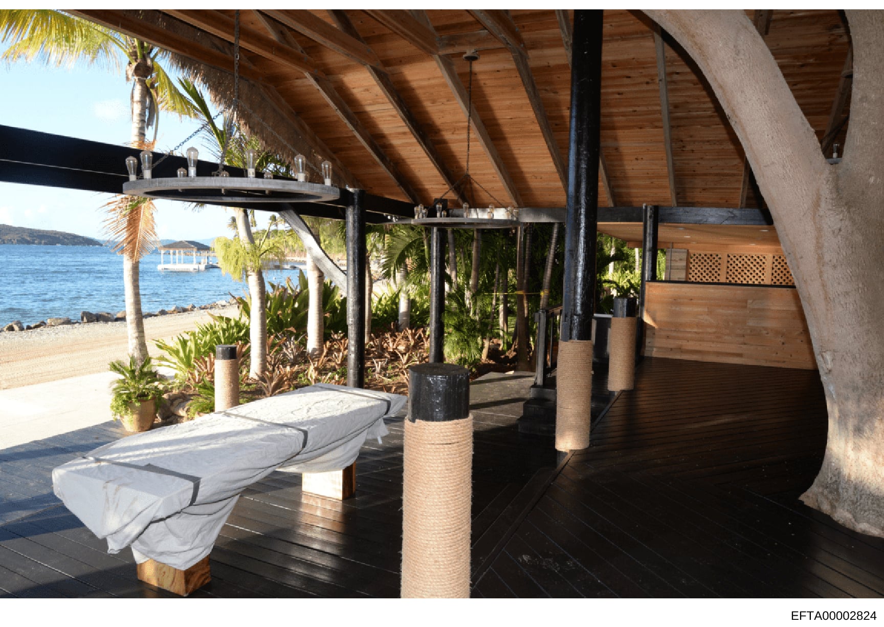 This photograph shows a luxury beachfront property's covered pavilion or outdoor living structure. The image depicts an open-air covered space with wooden ceiling beams, modern furnishings including loungers with cushions, decorative rope-wrapped col