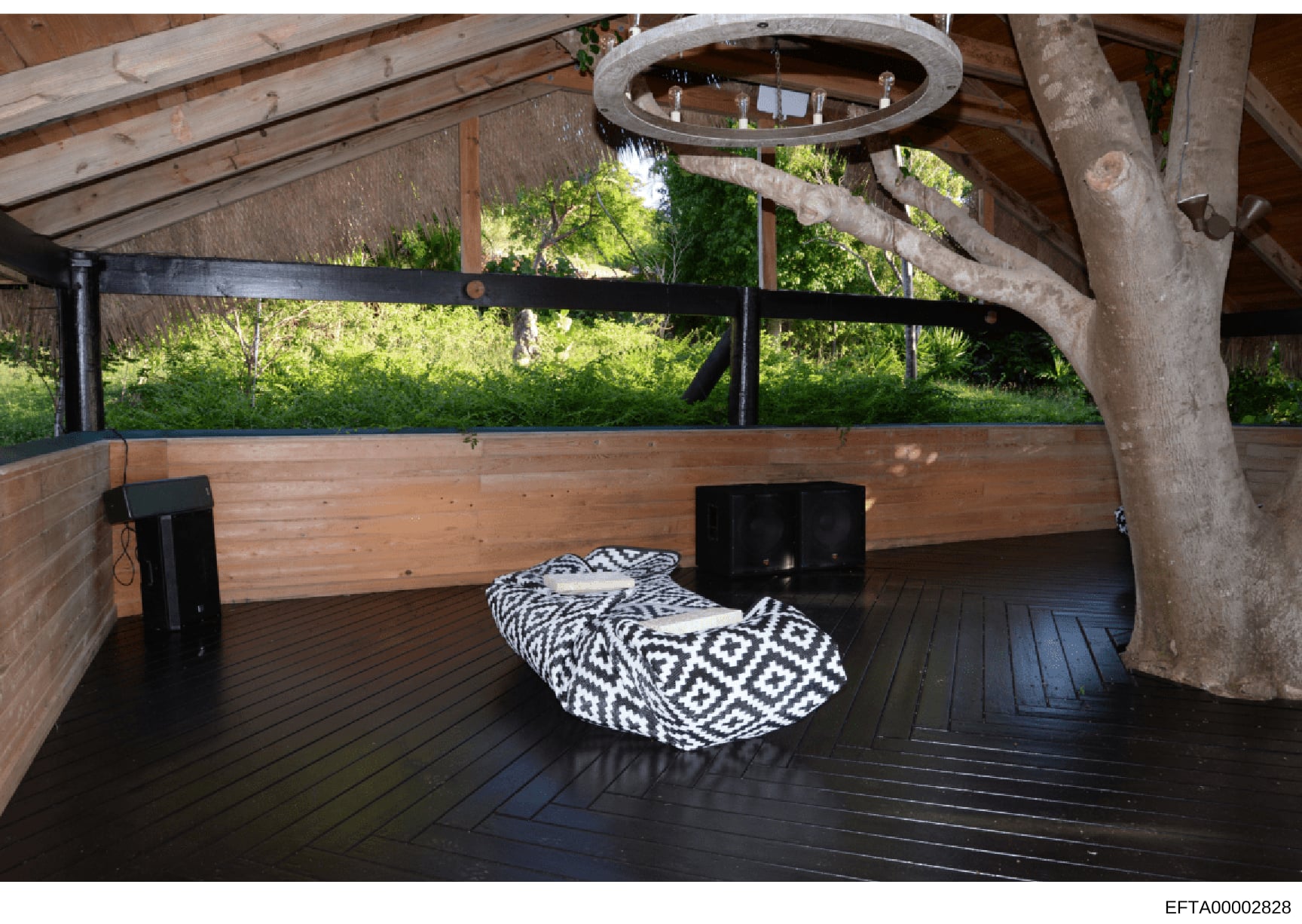 This photograph shows the interior of a covered outdoor structure with wooden decking, featuring rustic architectural elements including exposed beams and a large tree trunk. The space contains minimalist furnishings including black storage boxes and