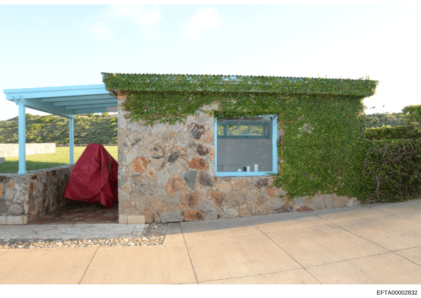 This is a photograph of a small stone cottage or outbuilding with distinctive blue metal trim, ivy-covered walls, and a covered patio area. The structure features a stone facade, blue-framed window and door, and ivy vines covering much of the upper p