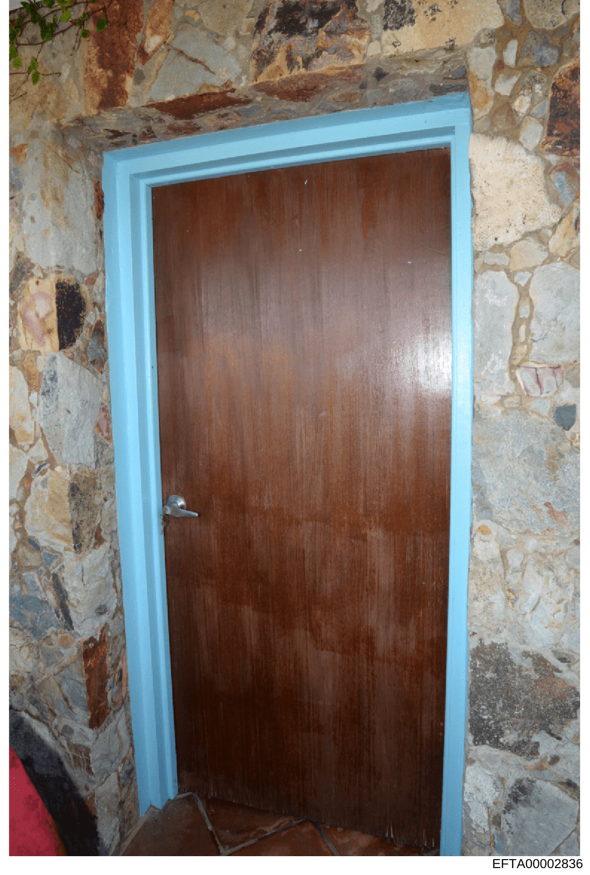 This is a photograph of an interior doorway featuring a dark wooden door with a bright blue frame set within weathered stone walls. The image appears to be documentation of a property's interior, possibly from one of Epstein's residences. The photogr