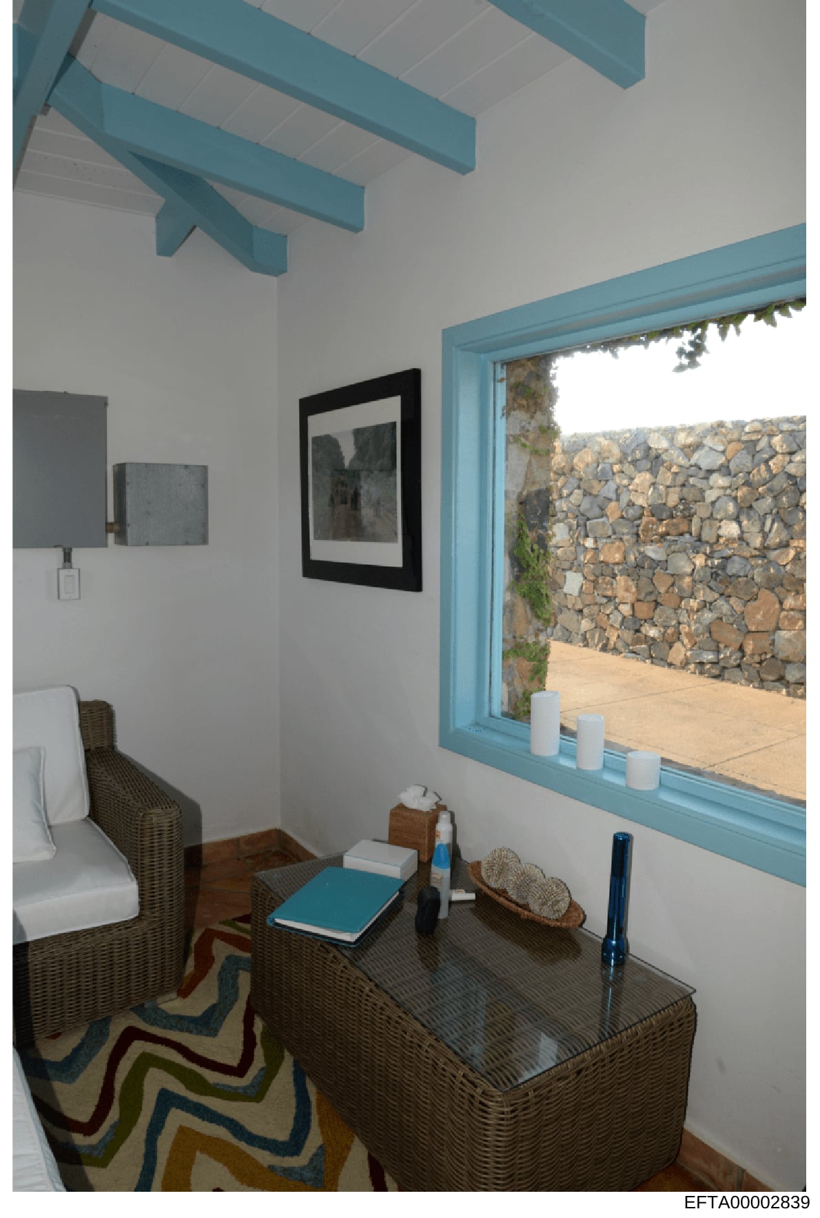 This photograph depicts an interior room, likely a bedroom or guest room, featuring modern design elements with turquoise architectural details and furnishings. The room has white walls, exposed turquoise ceiling beams, wicker furniture, and a large 