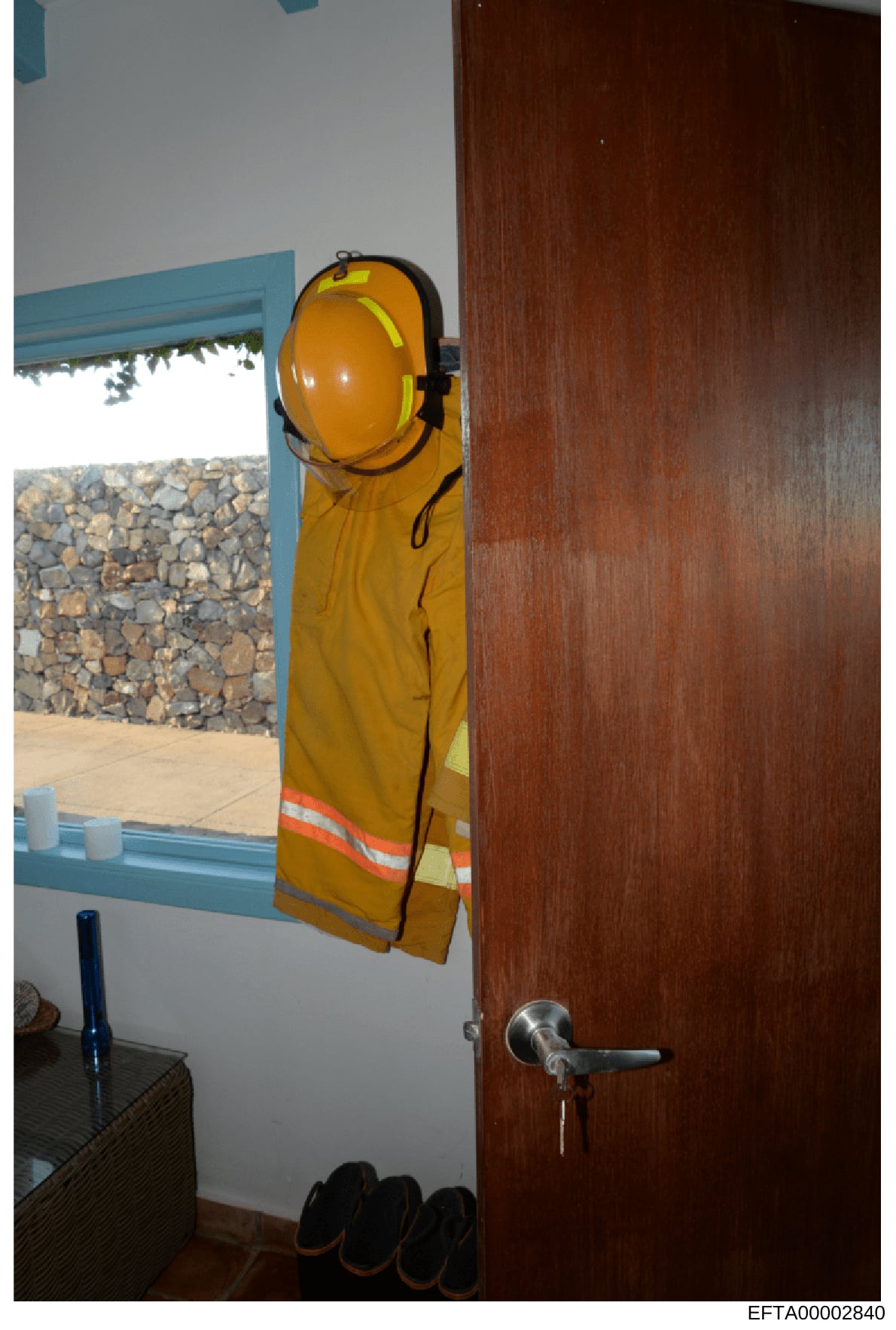 This is an evidence photograph showing the interior of a room with a window overlooking a stone courtyard or exterior area. A yellow hard hat and safety vest hang on the wall beside a dark wooden door. The image appears to be documentation of a prope