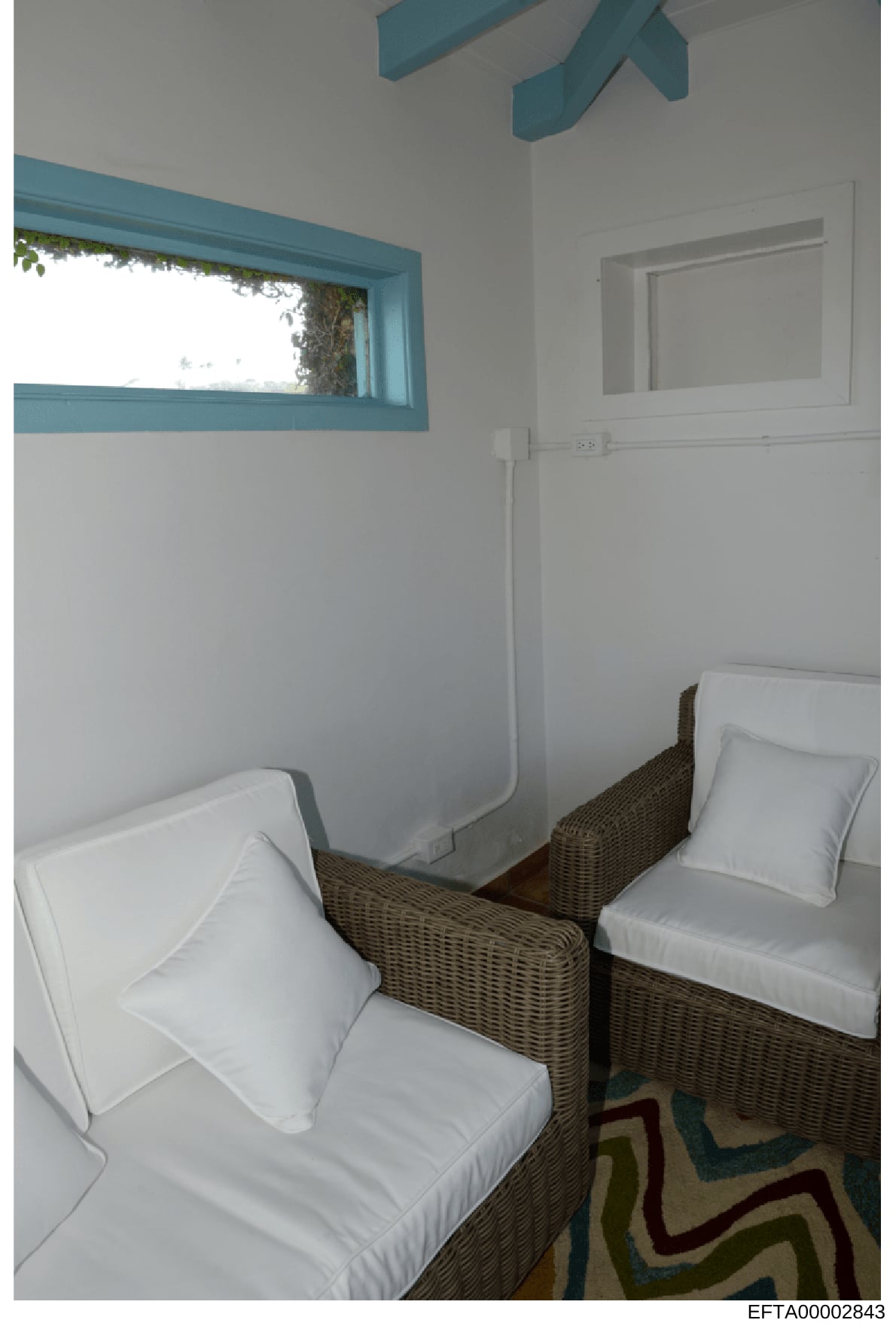 This is an evidence photograph showing a small bedroom interior with two twin beds with white linens and wicker headboards. The room features turquoise-trimmed windows and architectural elements, white walls, and minimal furnishings. The photograph a