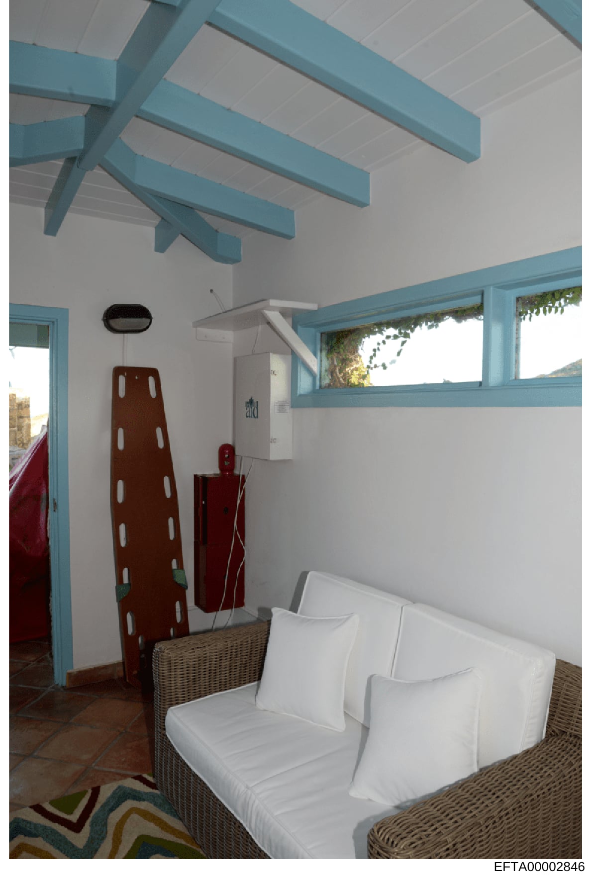 This is a photograph of an interior room featuring modern coastal-style design with turquoise and white color scheme. The room contains a wicker seating area with white cushions, exposed beam ceiling details, and large windows with ocean views. The i