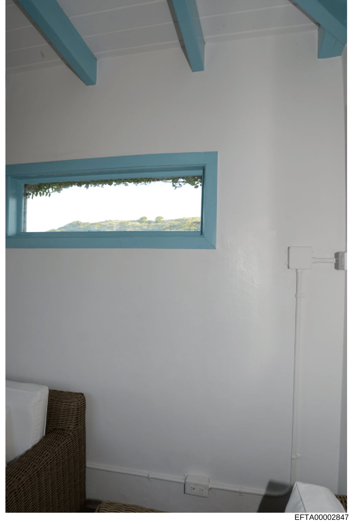 This is a photograph of an interior room, likely a bedroom or guest room, showing architectural details including turquoise-painted ceiling beams and window frames, white walls, and a horizontal window with a landscape view. The image appears to be e