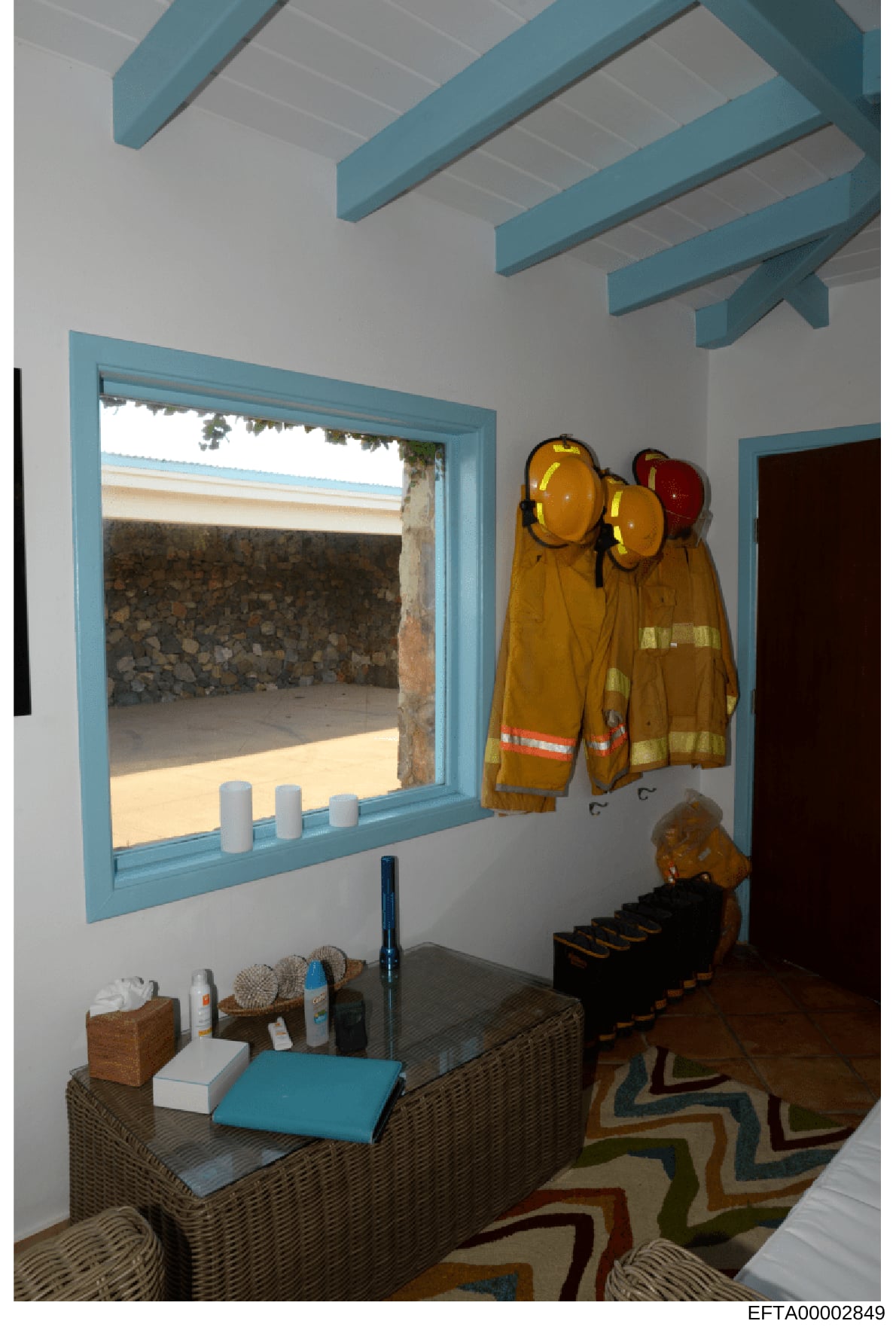This photograph shows an interior room with distinctive turquoise wooden ceiling beams and trim, furnished with a wicker console table beneath a large window. The room contains emergency/safety equipment including yellow firefighter jackets and hard