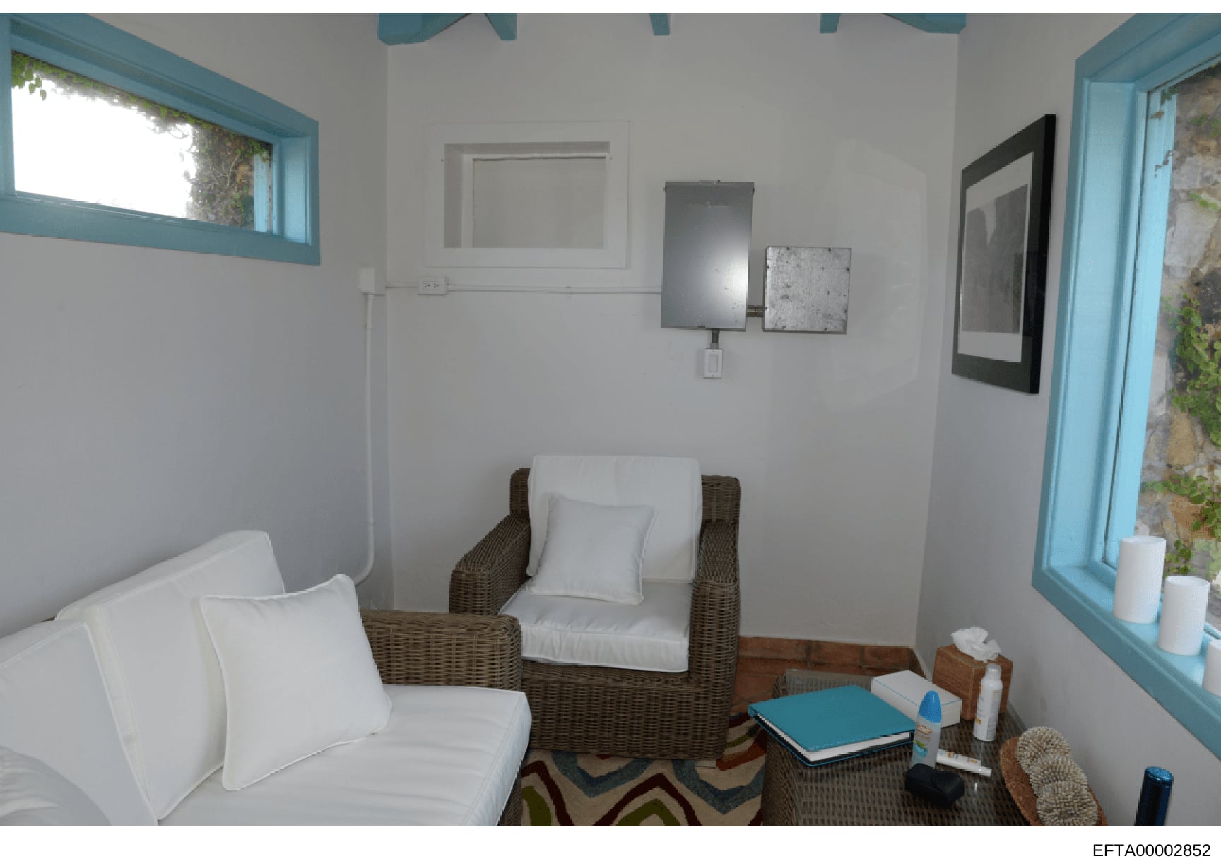 This is an interior photograph of a residential room featuring wicker seating furniture, white pillows, and basic furnishings. The room has turquoise-trimmed windows and doors, white walls, and appears to be a modest living or sitting area. The photo