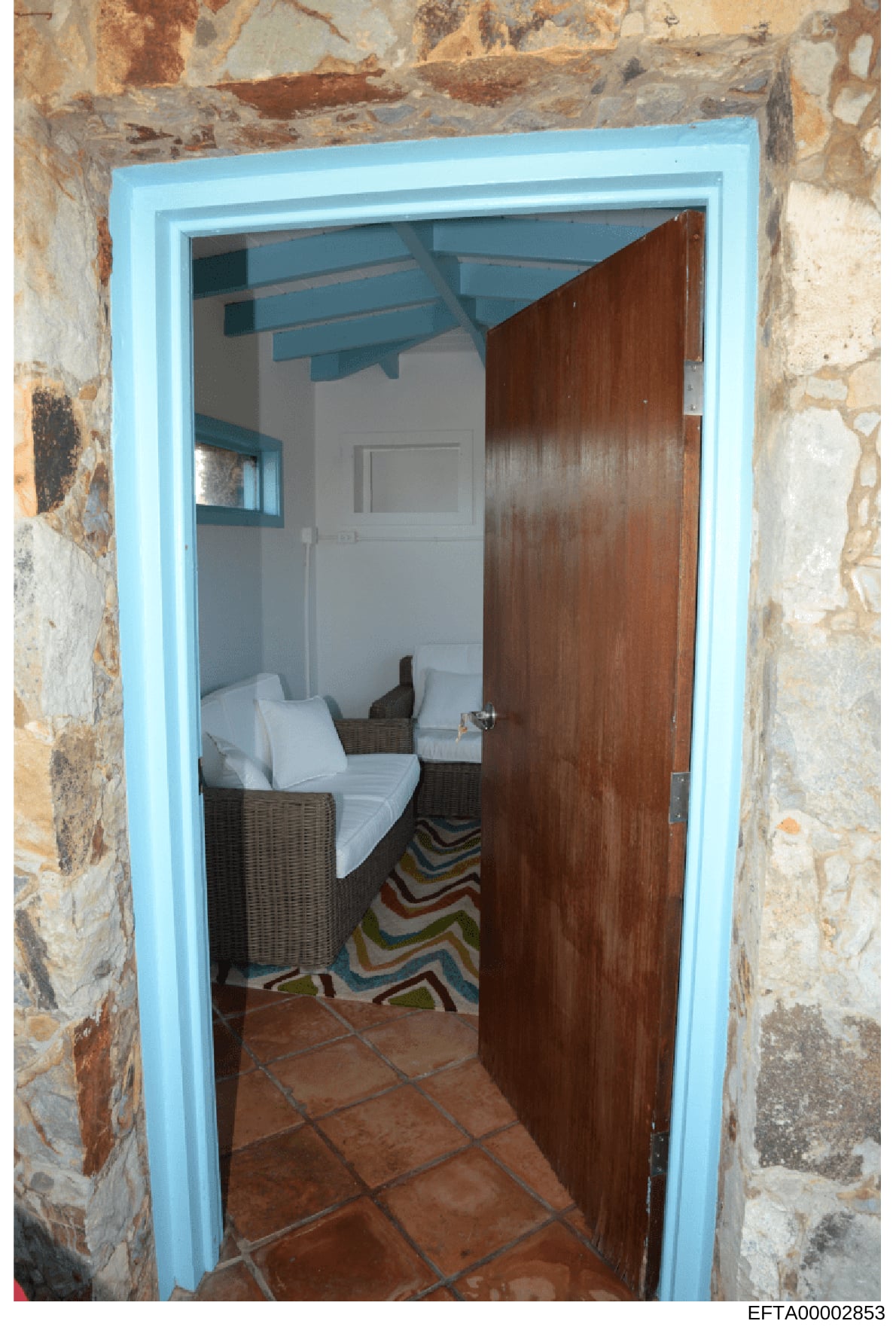 This is a photograph of an interior room, likely a bedroom or guest room, showing a doorway framed by weathered stone walls with a bright blue trim. The room contains wicker furniture with white cushions and a colorful striped rug. The image appears 