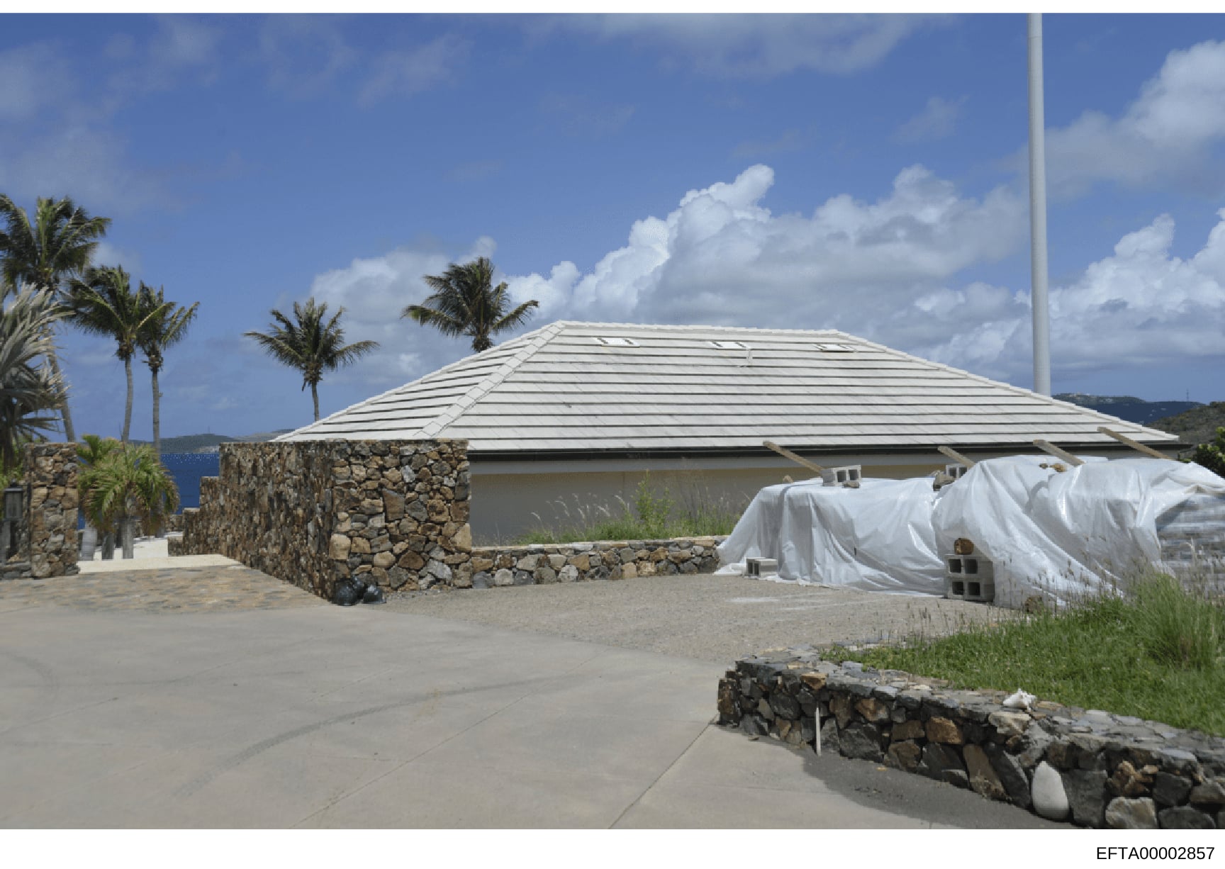 Photograph of a residential property under construction in a tropical coastal location, featuring stone masonry walls, a metal-paneled roof structure, and white protective sheeting covering portions of the building. The property appears to be situate