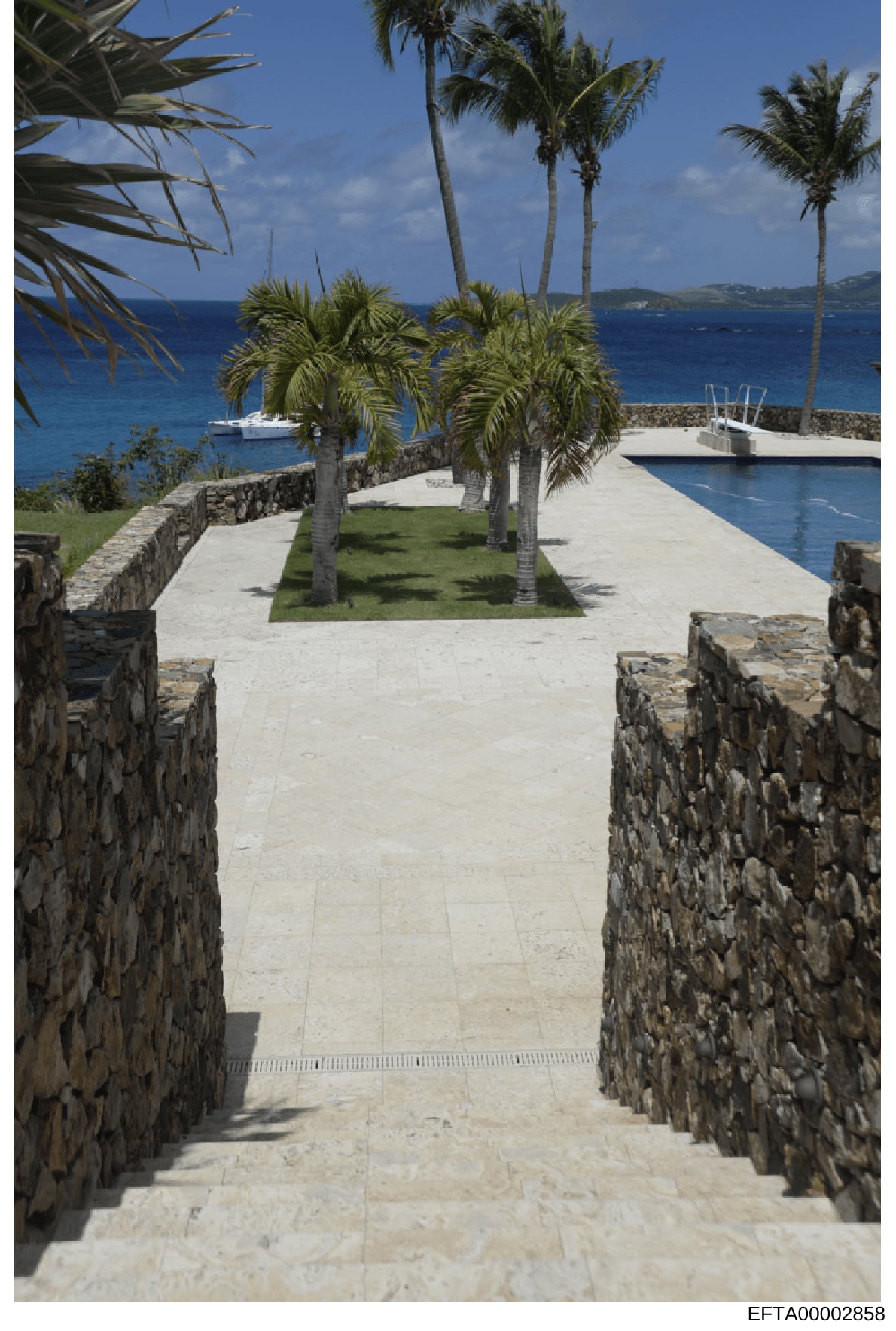 This photograph shows a luxurious waterfront property featuring a manicured courtyard with palm trees, an infinity pool, and a clear view of the turquoise Caribbean Sea with moored vessels. The stone-walled pathways and high-end construction suggest 