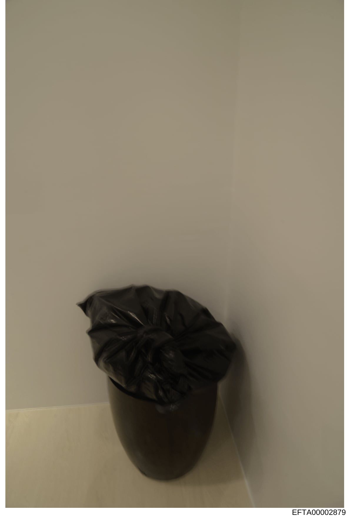 This is an evidence photograph showing a black plastic trash bag or similar container positioned against a white wall. The image appears to be part of the Epstein investigation documentation, with a Bates stamp indicating it was cataloged as evidence