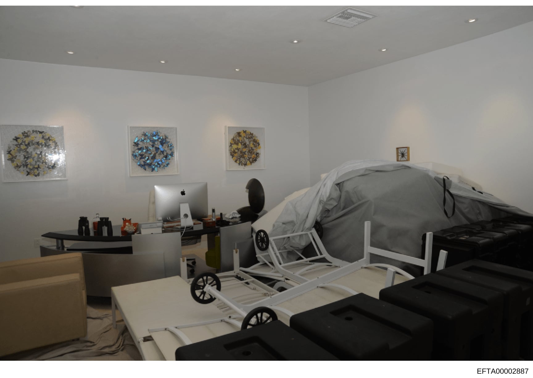 This is a photograph of an interior room, likely part of an Epstein property, showing a modern office space with an Apple iMac computer workstation, wall-mounted artwork, and what appears to be covered furniture or equipment. The room features recess