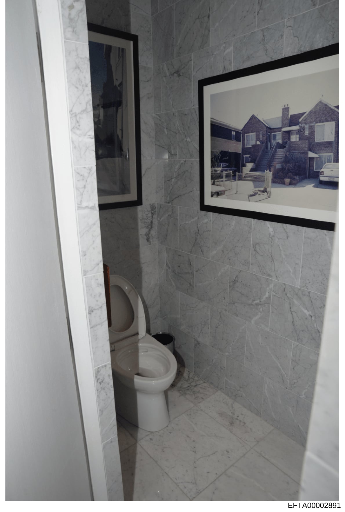 This is a photograph of a bathroom interior, likely from one of Jeffrey Epstein's properties, showing marble tile walls, a white toilet, and a framed black-and-white photograph of a residential building mounted on the wall. The image was collected as