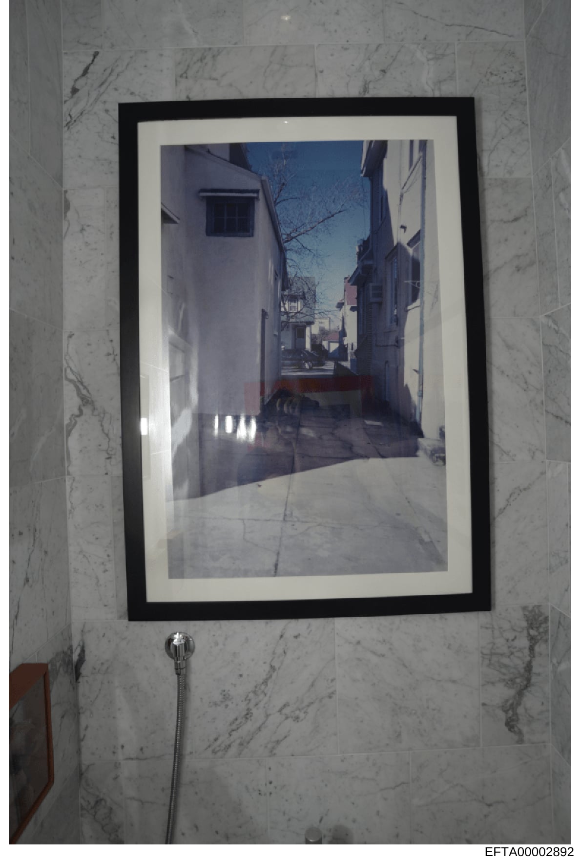 This is a photograph of a framed artwork depicting a narrow European street with white-washed buildings, bare trees, and clear blue sky. The photograph shows the framed print hanging on a marble-tiled wall in what appears to be a bathroom, with a sho