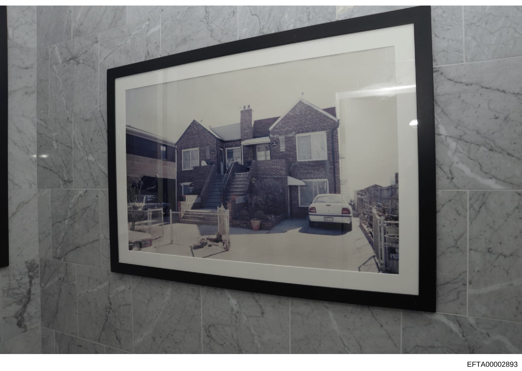 This is a photograph of a framed image showing the exterior of a brick residential property with multiple chimneys, external stairs, windows, and a parked vehicle in the driveway. The framed photograph is mounted on a marble-tiled wall. The image app