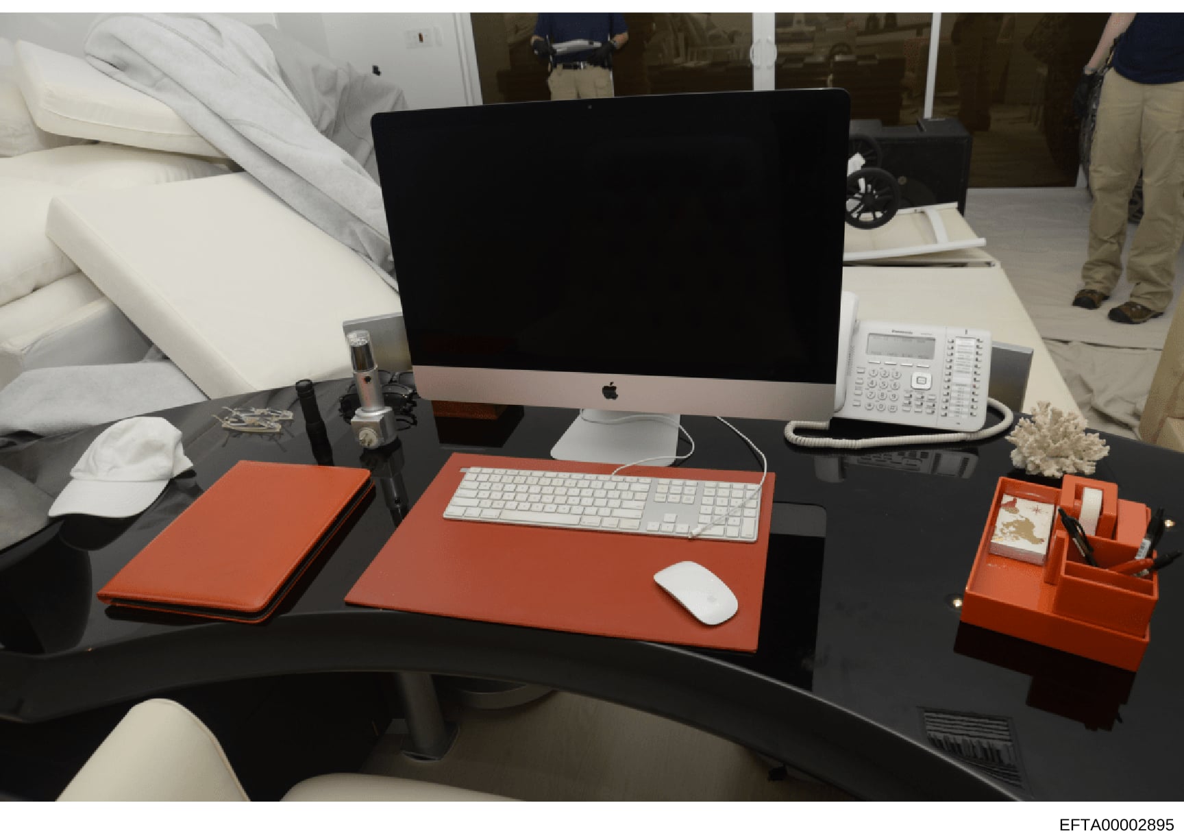 This photograph shows a professional office desk arrangement featuring a modern Apple iMac computer, white keyboard and mouse, and various orange-colored desk accessories including a mousepad, organizer, and notebook. The desk also includes a white t