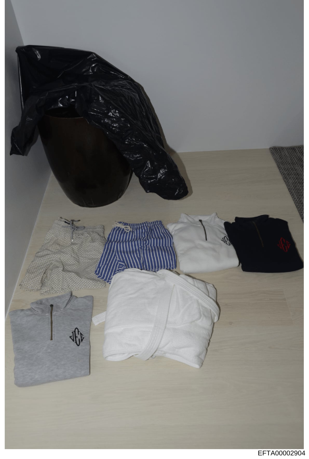 This is an evidence photograph showing clothing items laid out on a light-colored surface, including underwear, striped shorts, t-shirts with monograms, and a black plastic bag. The items appear to be catalogued evidence from a search or seizure cond