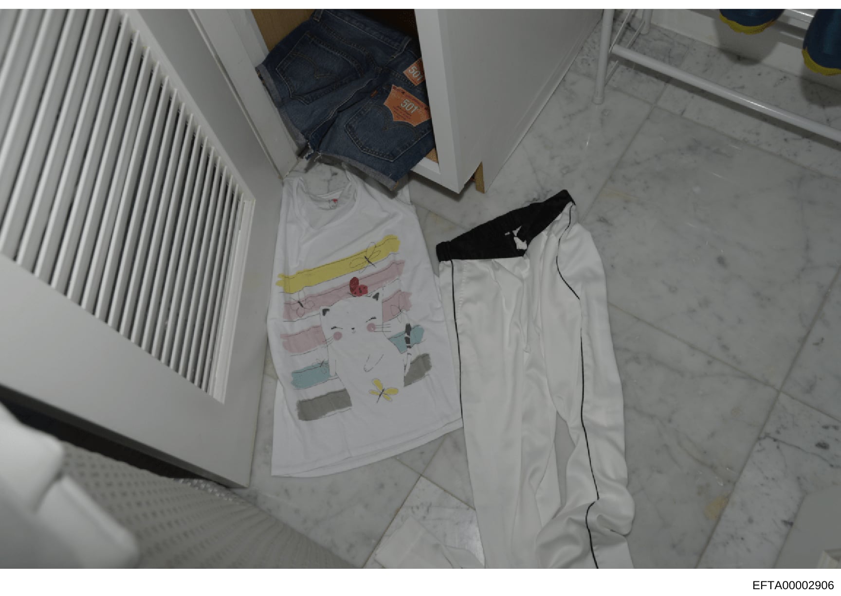 This is an overhead photograph of clothing and personal items laid out on a marble floor, including jeans, a white shirt with colored drawings, white pants, and a black garment. The photo appears to be evidence documentation from an investigation, sh