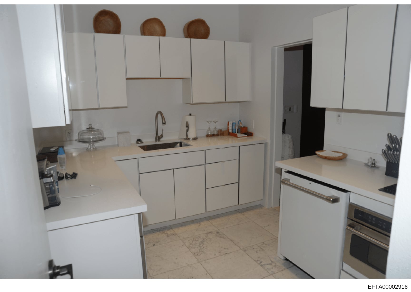 This is a photograph of a modern kitchen interior, showing white cabinetry, countertops, appliances, and fixtures. The image appears to be part of property documentation or evidence collection related to an Epstein-connected property. The photo is da