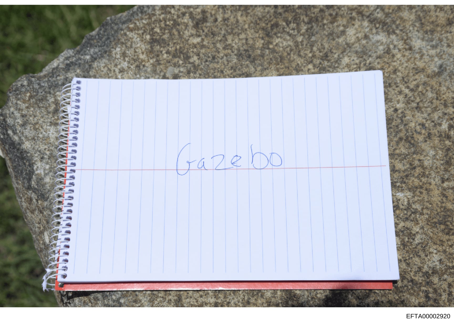 This photograph shows a spiral-bound notebook page with the word 'gazebo' written in blue pen. The notebook is positioned on what appears to be a concrete or stone surface outdoors. The minimal text and casual nature of the document suggest it may be