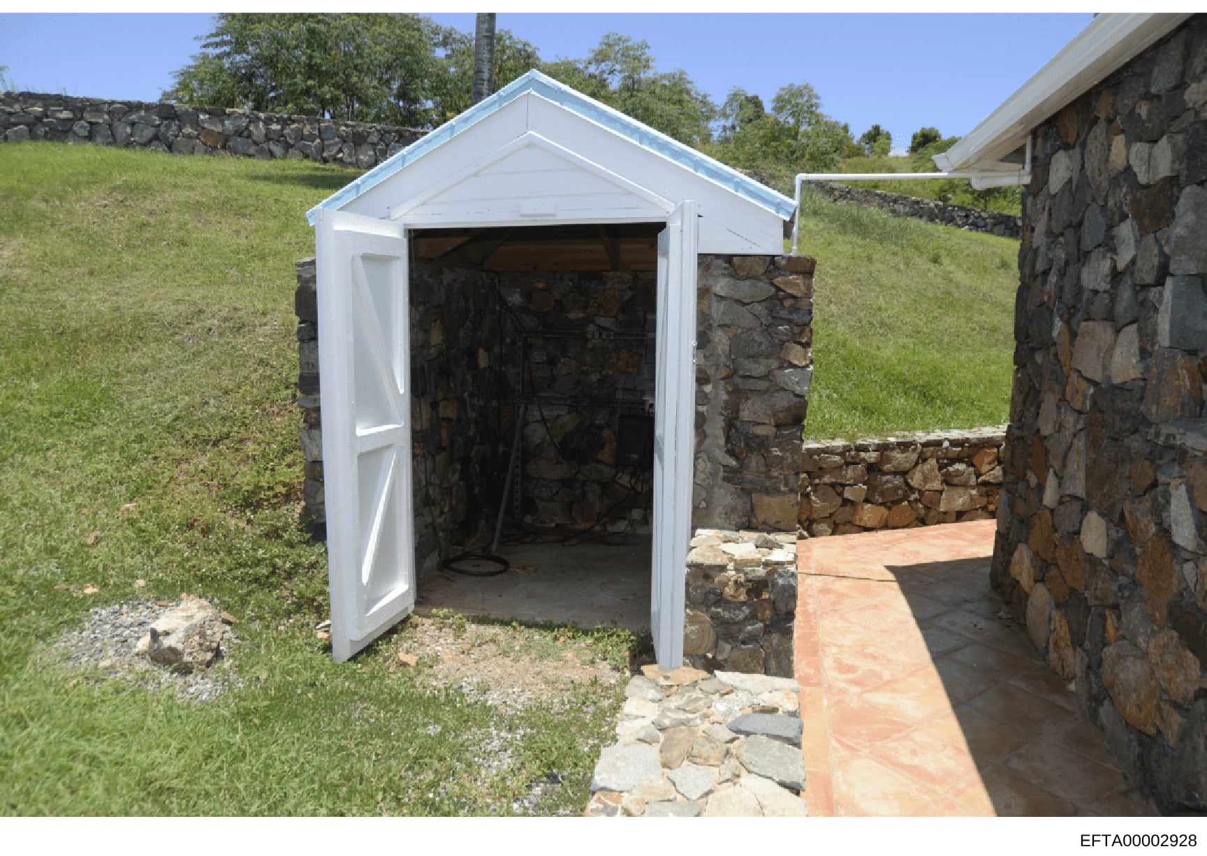 This is a photograph of a white-painted storage shed or outbuilding on what appears to be an estate property. The structure features a gabled roof, open double doors, and is built partially into or adjacent to existing stone foundation walls. The set