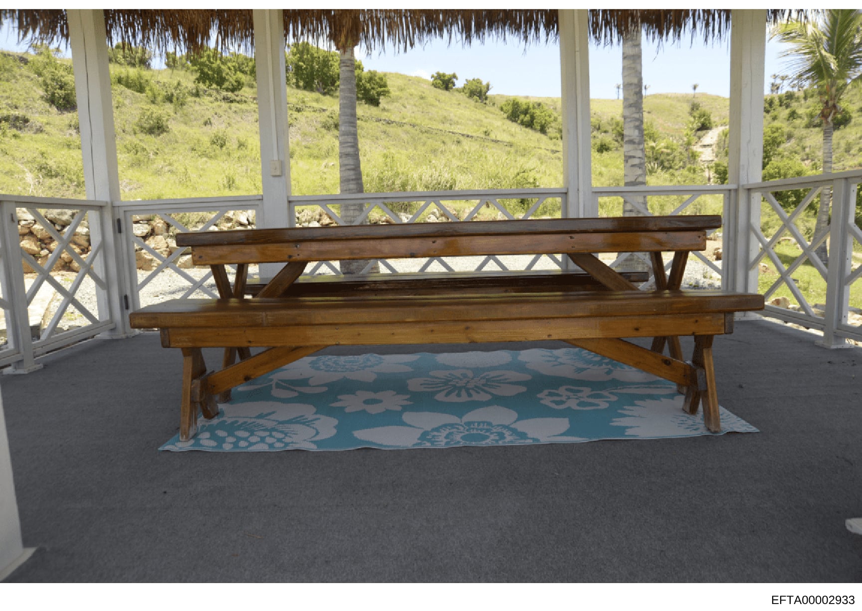 This is a photograph of an interior patio or covered gazebo area featuring a wooden bench positioned on a decorative blue and white patterned rug. The space overlooks lush, green hillside terrain with palm trees visible through screened windows and d