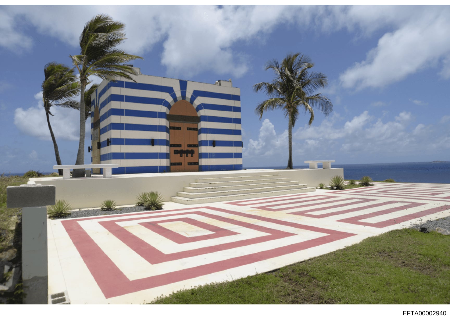 This is a photograph of a unique oceanfront property featuring a striking modernist building with blue and white horizontal stripes and a large geometric wooden entrance door. The property includes manicured grounds with red and white geometric pavin