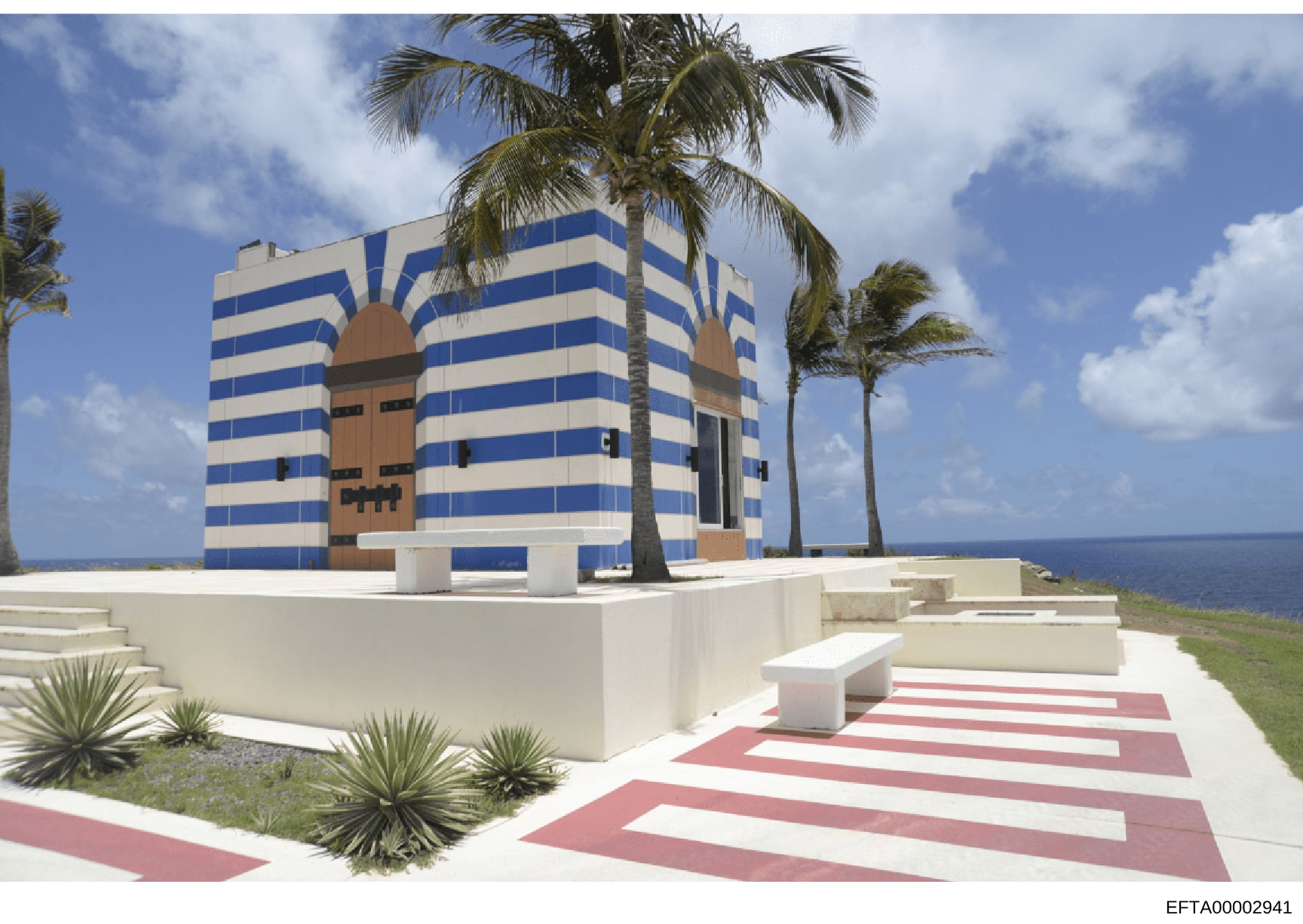 This is a photograph of a distinctive modern building with bold blue and white horizontal stripes and a stylized brown central design element, located on an oceanfront property. The structure features palm trees, manicured landscaping, and a striped