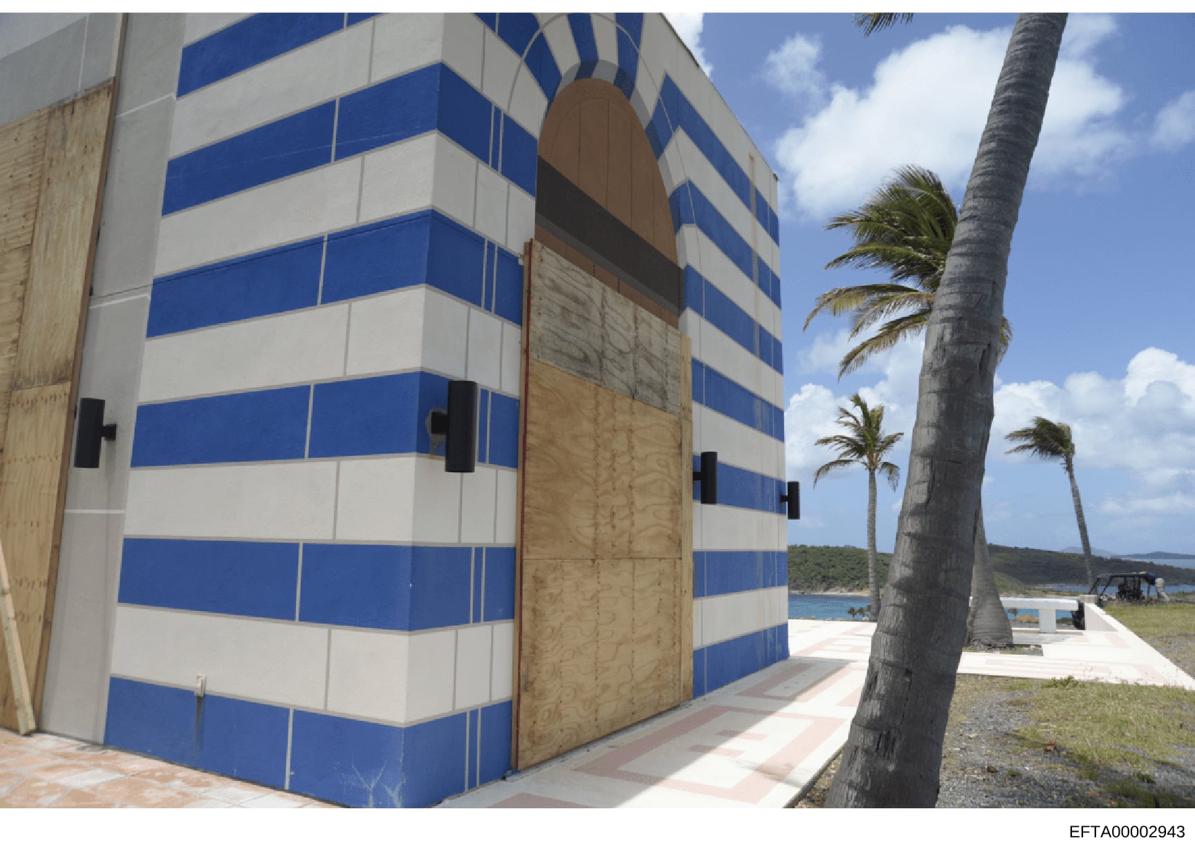 This photograph shows a distinctive blue and white striped residential or commercial building in a tropical coastal setting. The building features horizontal blue and white stripes, wooden doors, and is situated near a beach with palm trees visible. 