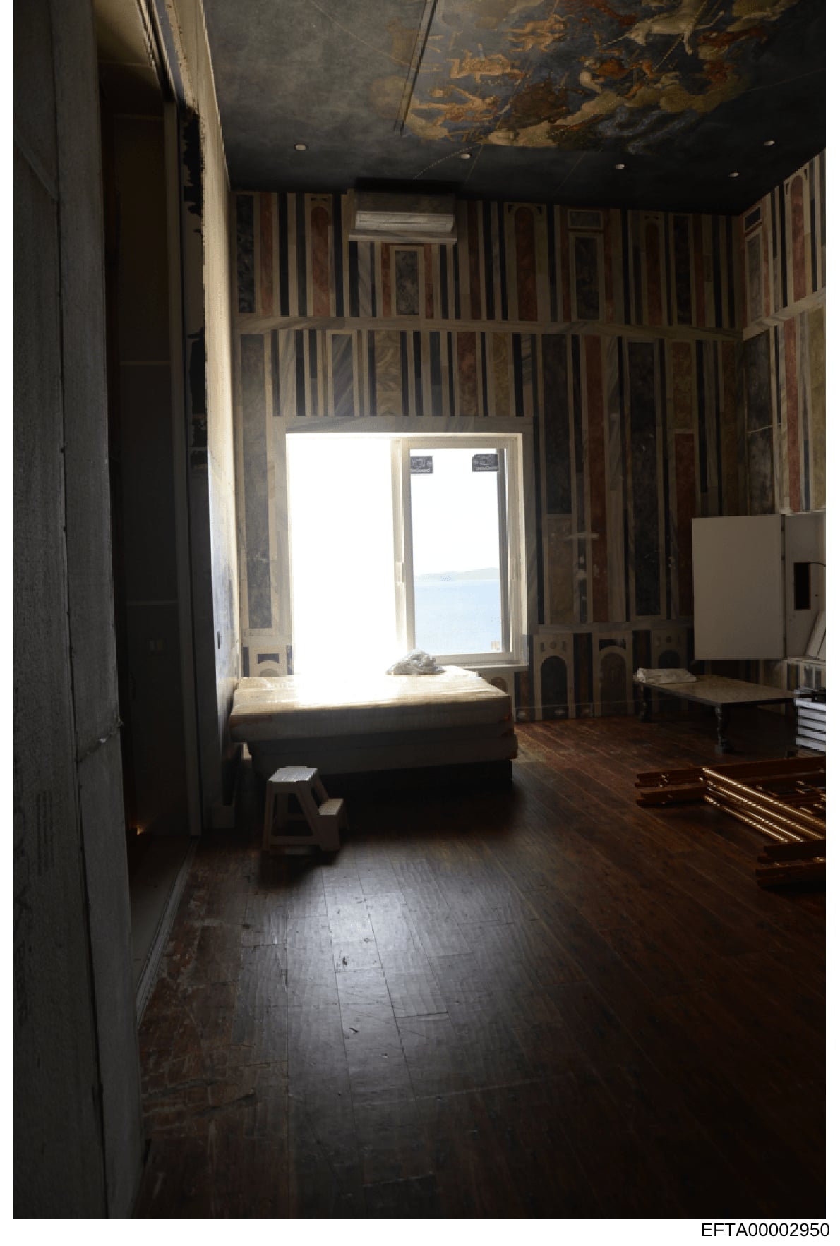 This is a photograph of an interior room, likely a bedroom or living space, showing ornate striped wall paneling, a dark ceiling with decorative details, wooden flooring, and large windows offering a view of water. The room appears to be part of a re