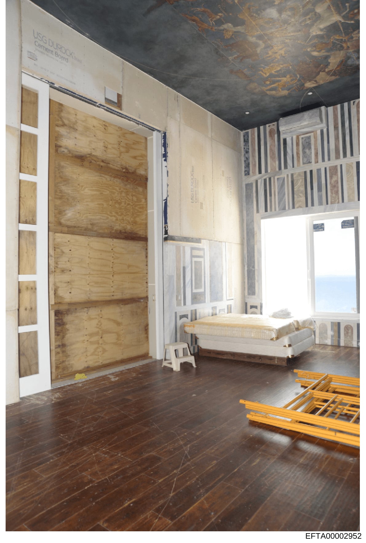 This is an evidence photograph showing the interior of a bedroom during renovation or construction. The room features boarded-up windows and doors, exposed construction materials, and ocean views visible through open areas. The photo documents the st