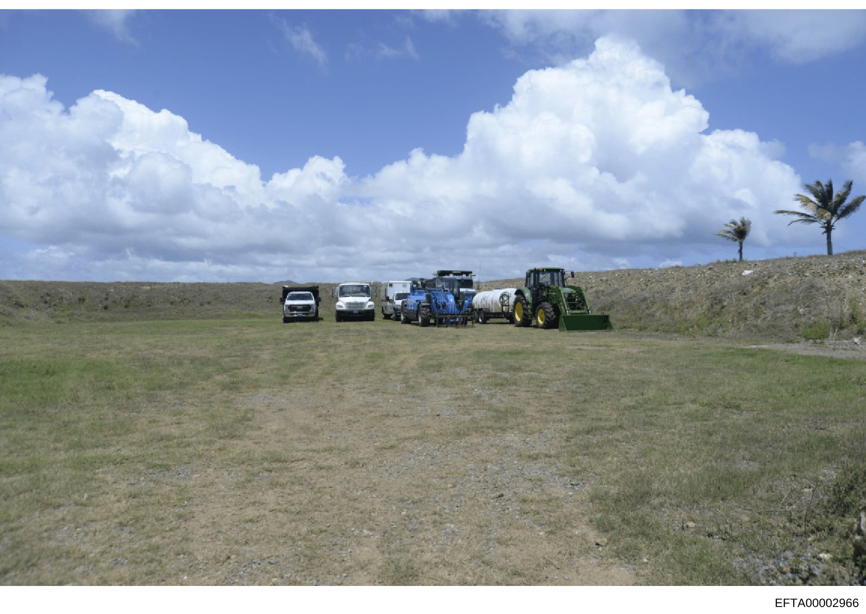 This photograph shows a rural property or ranch with multiple vehicles and agricultural equipment lined up in an open field. The image depicts what appears to be a working ranch or agricultural facility with tractors, trucks, and other heavy equipmen