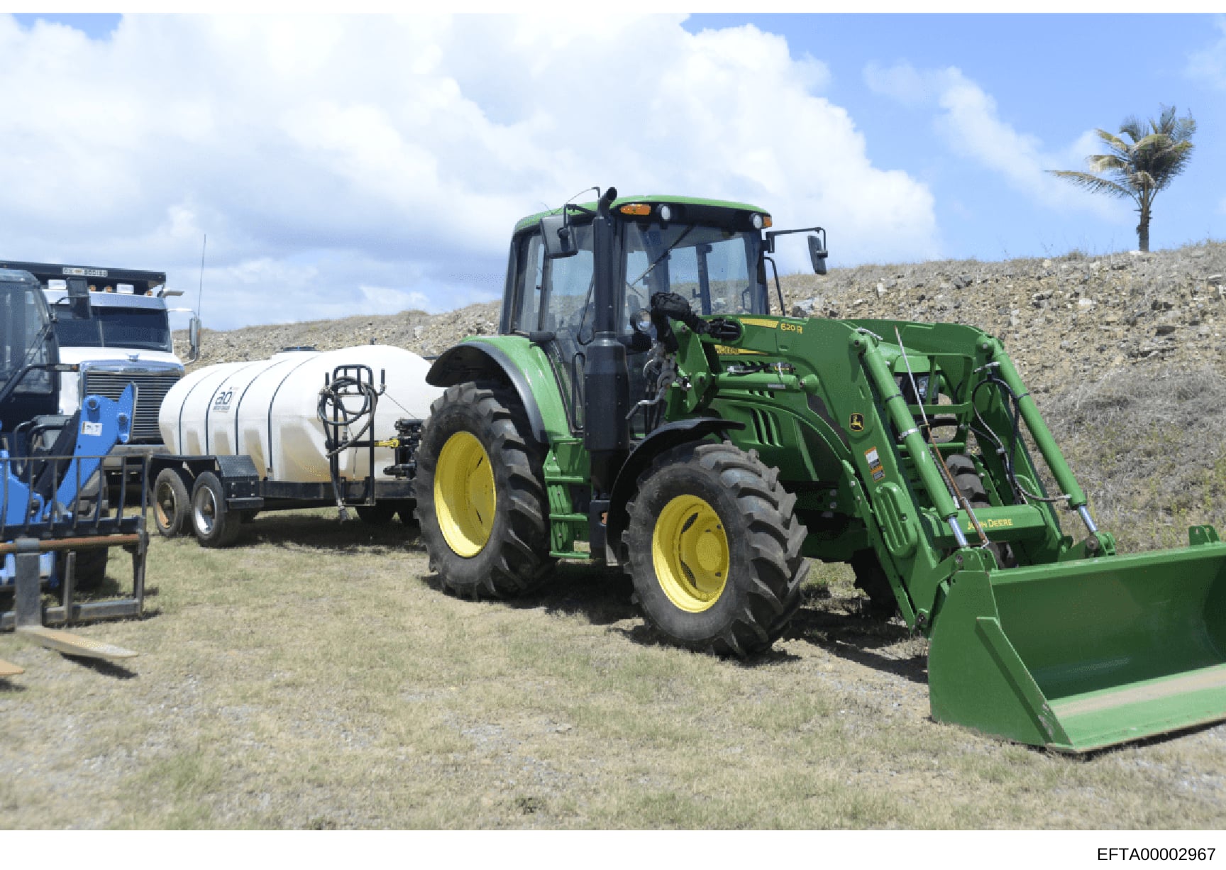 This is a photograph showing agricultural and heavy equipment at what appears to be a rural or property maintenance site. The image depicts a green John Deere tractor with front-end loader, a white water/liquid transport tank trailer, and other suppo