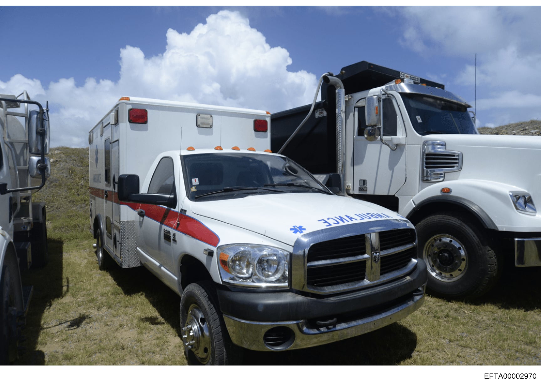 This photograph shows three emergency medical service (EMS) vehicles parked together in an outdoor, grassy area with hills in the background. The image displays a white ambulance with red and blue markings in the foreground, a white ambulance unit in