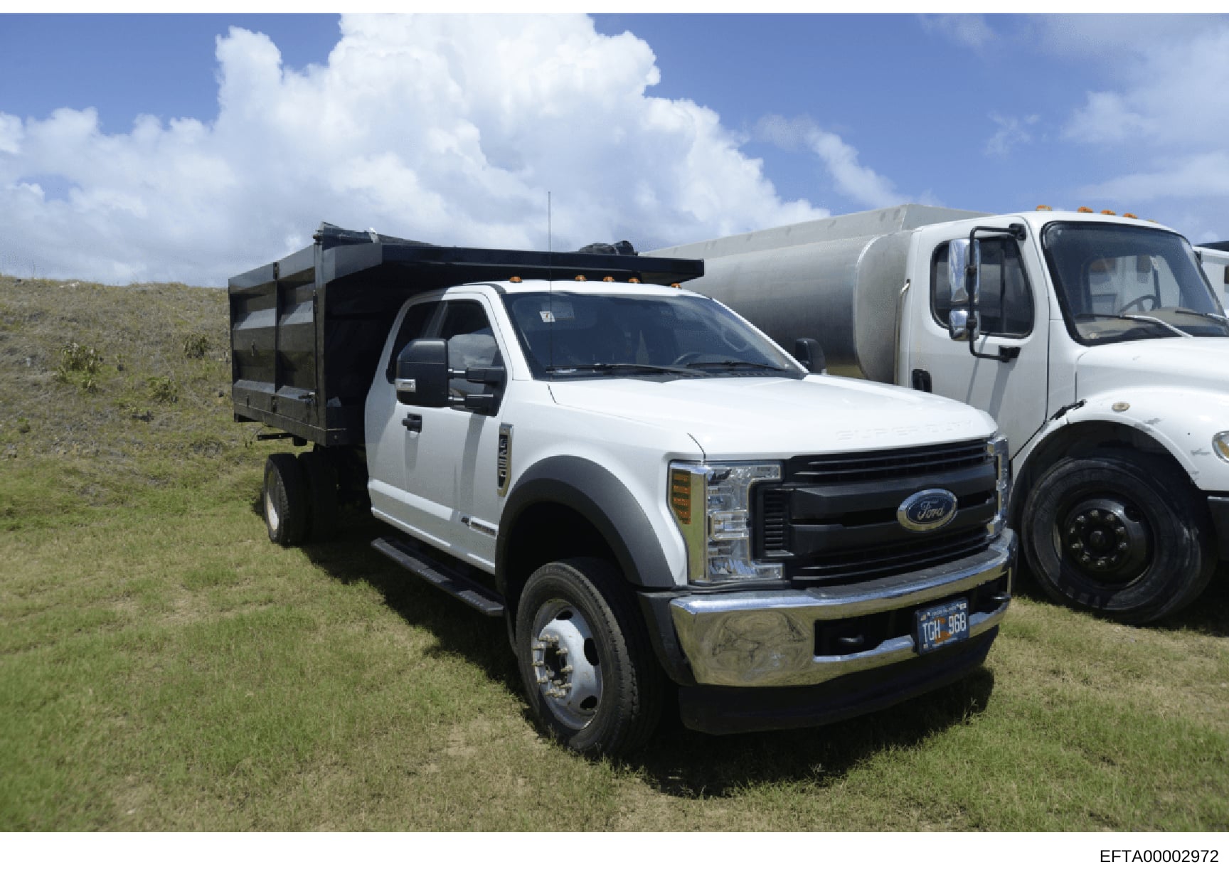 Photograph showing commercial vehicles parked on a grassy property, including a white Ford dump truck with black bed and a white tanker/transport truck. The image appears to be documentation of equipment or vehicles associated with a property or faci