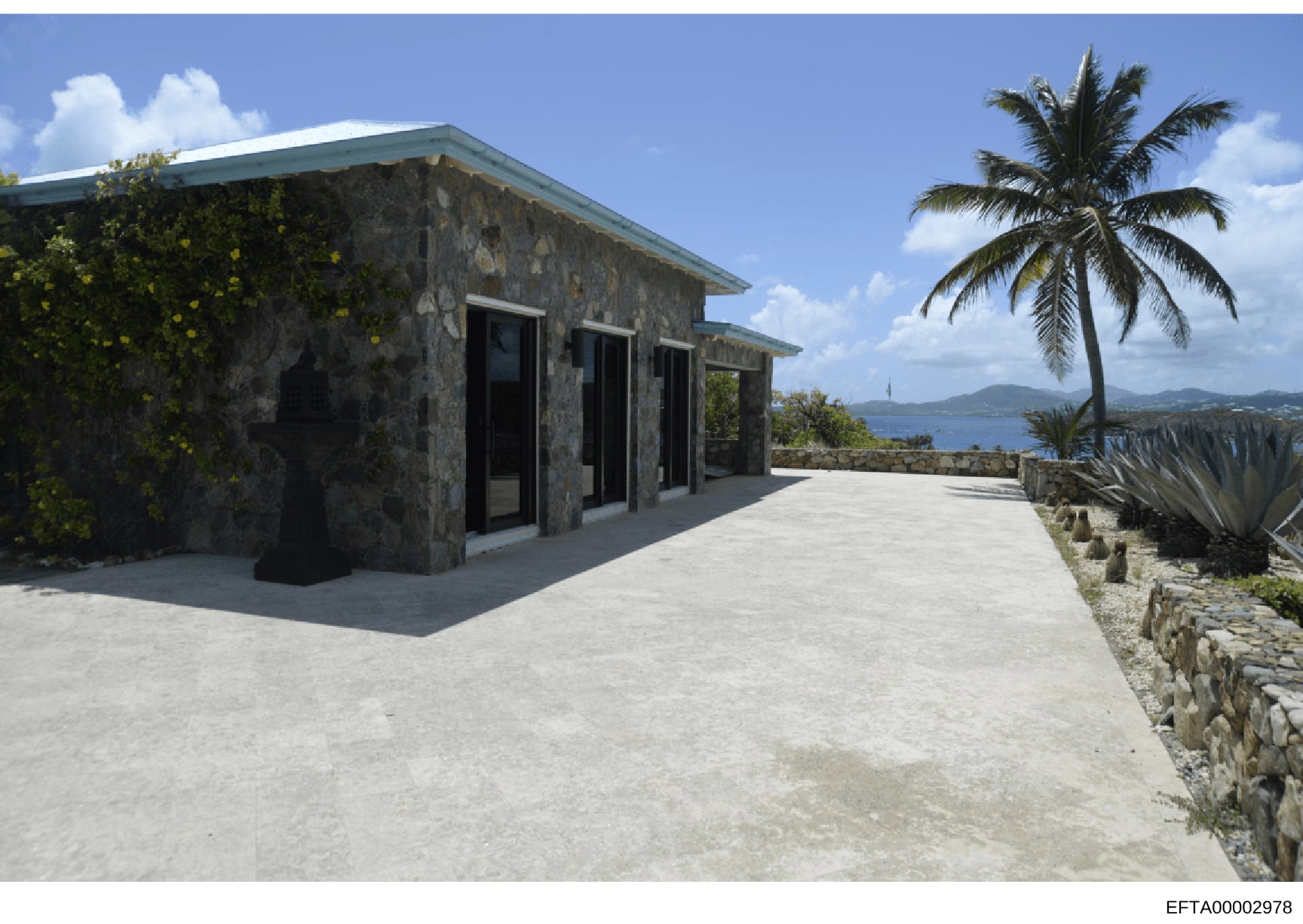 This is a photograph of a modern stone structure located on a coastal hillside with panoramic ocean views. The building features contemporary architecture with a turquoise-trimmed roof, large glass doors/windows, and an expansive concrete terrace. Th
