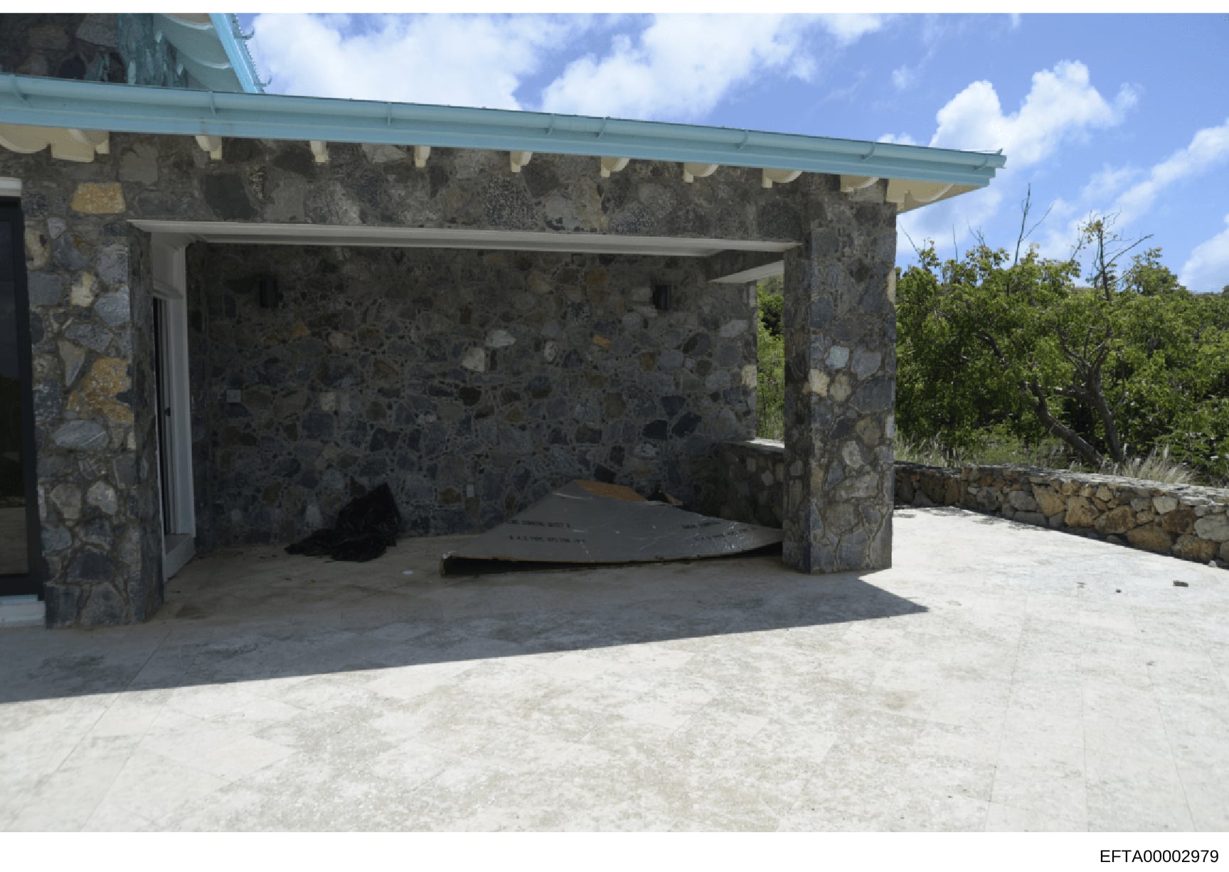 This is a photograph of an exterior structure featuring stone construction with a turquoise metal roof overhang. The building appears to be a pavilion or outbuilding with an open front design, situated in a tropical or subtropical location with veget