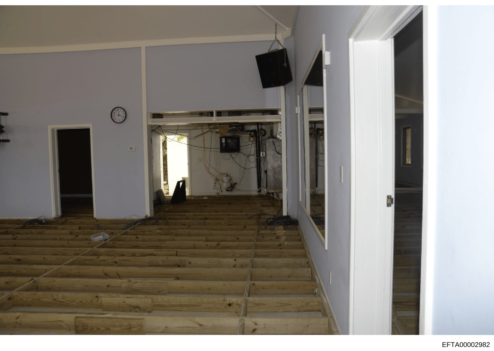 This photograph shows an interior room or commercial space that appears to be undergoing renovation or construction. The space features light blue/lavender walls, wooden flooring, multiple doorways, exposed electrical wiring, and construction equipme