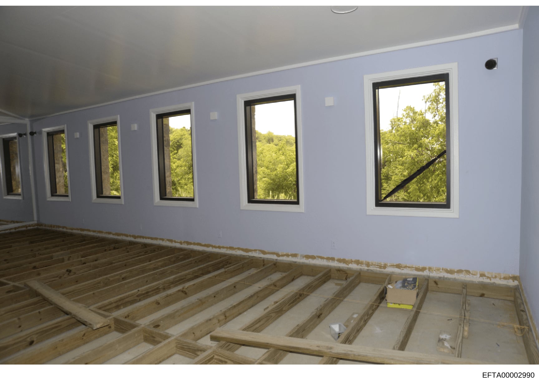 This is a photograph of an interior room under construction or renovation, featuring light blue-painted walls, multiple large black-framed windows overlooking wooded landscape, and exposed wooden floor joists. The image appears to be documentation of
