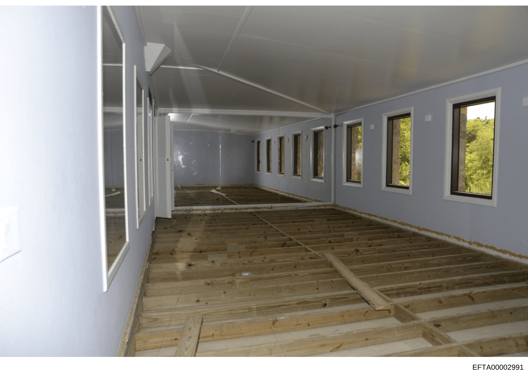 This photograph documents the interior of a room during construction or renovation, featuring light blue/gray walls, multiple windows with views of greenery, wooden subfloor framing, and white trim elements. The image appears to be part of property d