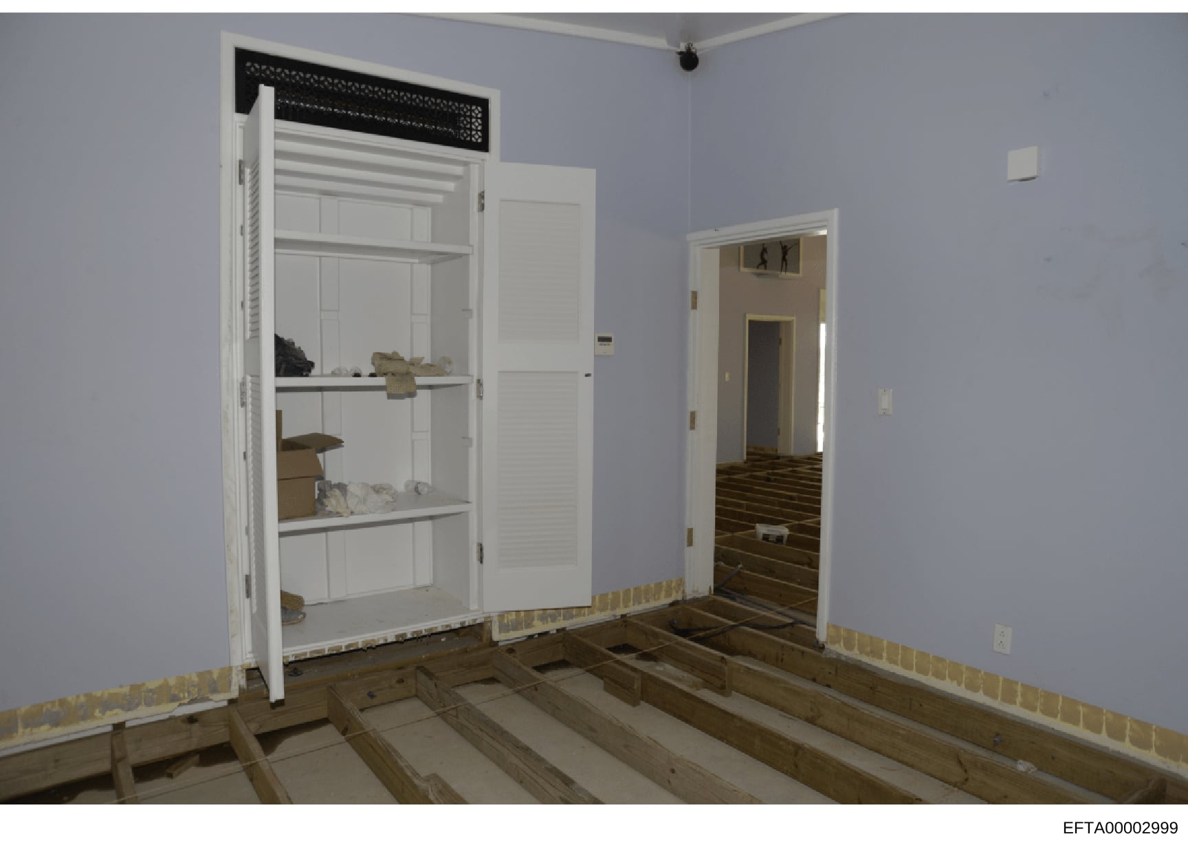This is a photograph of an interior room in an early stage of renovation or construction. The space features exposed wooden floor joists, light blue/periwinkle walls, white trim and doors, and built-in shelving with stored items. The image appears to
