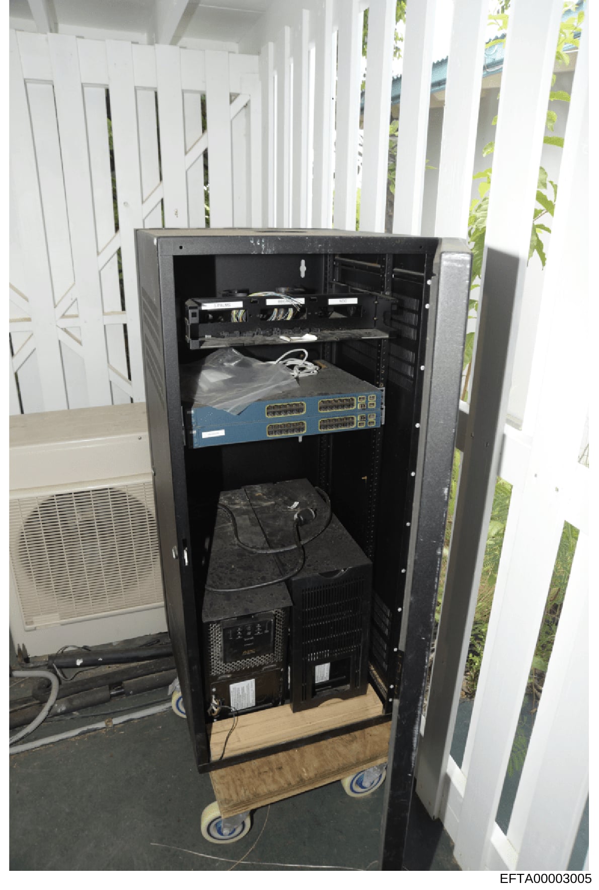 This photograph shows a black server rack containing networking equipment, including a blue network switch with multiple ports, cabling, and power distribution hardware. The rack is positioned in what appears to be an outdoor or semi-enclosed white s