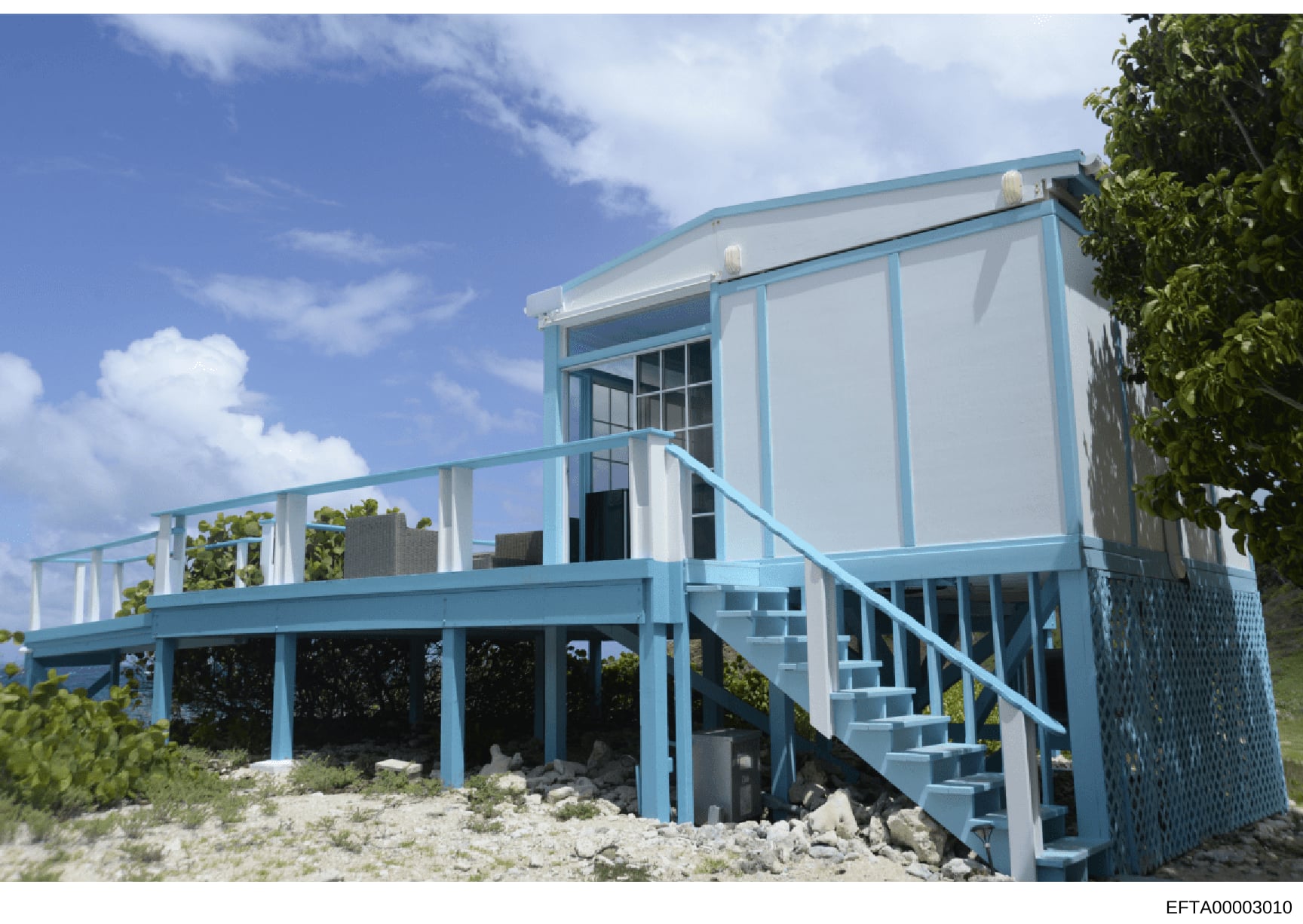 This is a photograph of a modern beachfront residential structure featuring blue and white exterior paneling, elevated on stilts above sandy terrain. The building appears to be a contemporary beach house or property with an open-air deck, multiple le